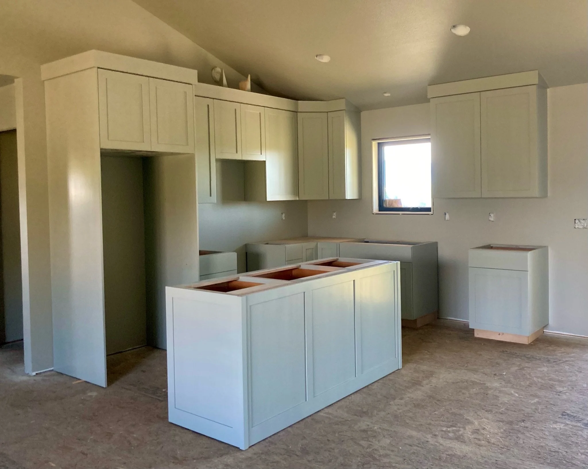 A kitchen under construction with light-colored cabinets, some open compartments, a window, and a wooden floor, with exposed wall outlets and no appliances installed yet.