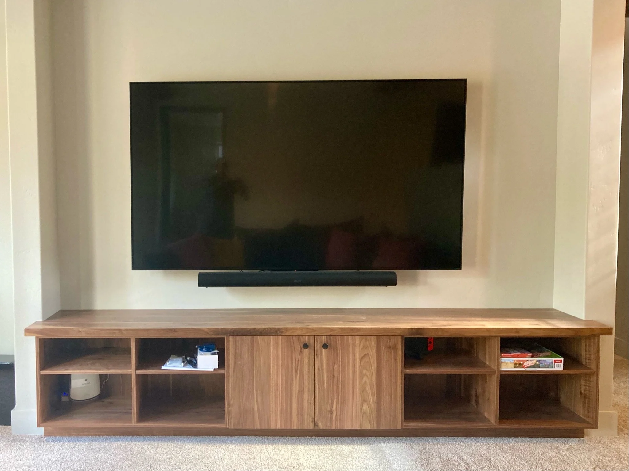 A flat-screen television mounted on a beige wall above a wooden entertainment center with open shelves, containing books, electronic devices, and decorative items.