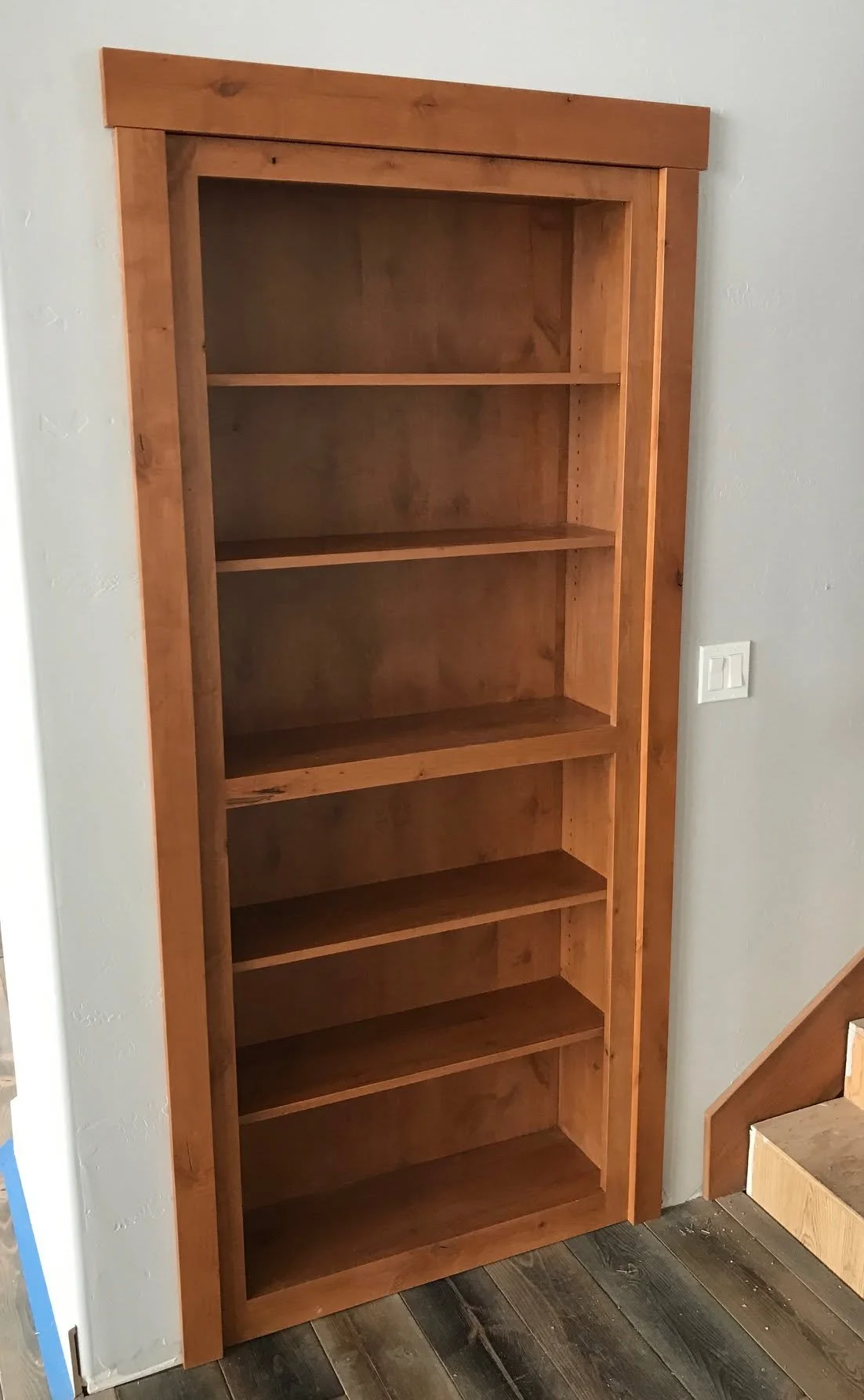 Wooden bookshelf with five shelves positioned against a wall near stairs.