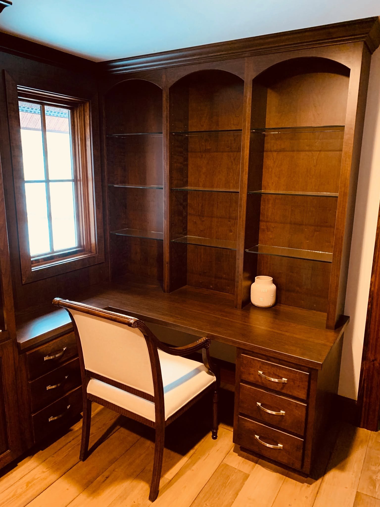 A wooden built-in bookshelf with empty shelves, next to a window, with a white ceramic vase on the desk in front of it.