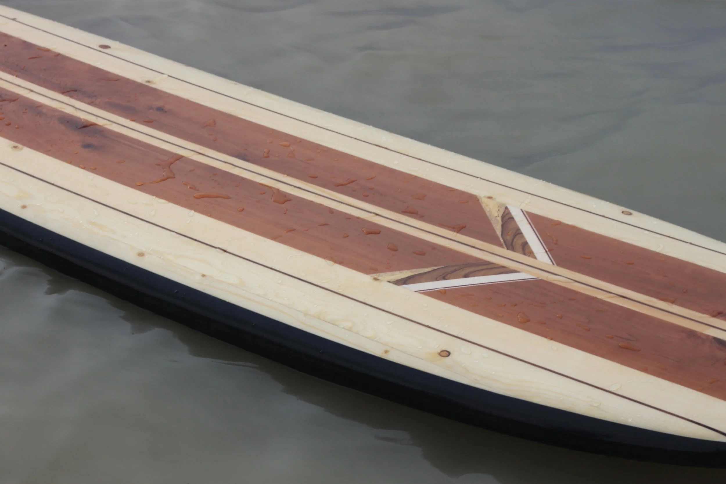 A close-up of a boat deck made of wood, with wet spots and water droplets on its surface. The deck has white and reddish-brown planks. The water in the background is calm.