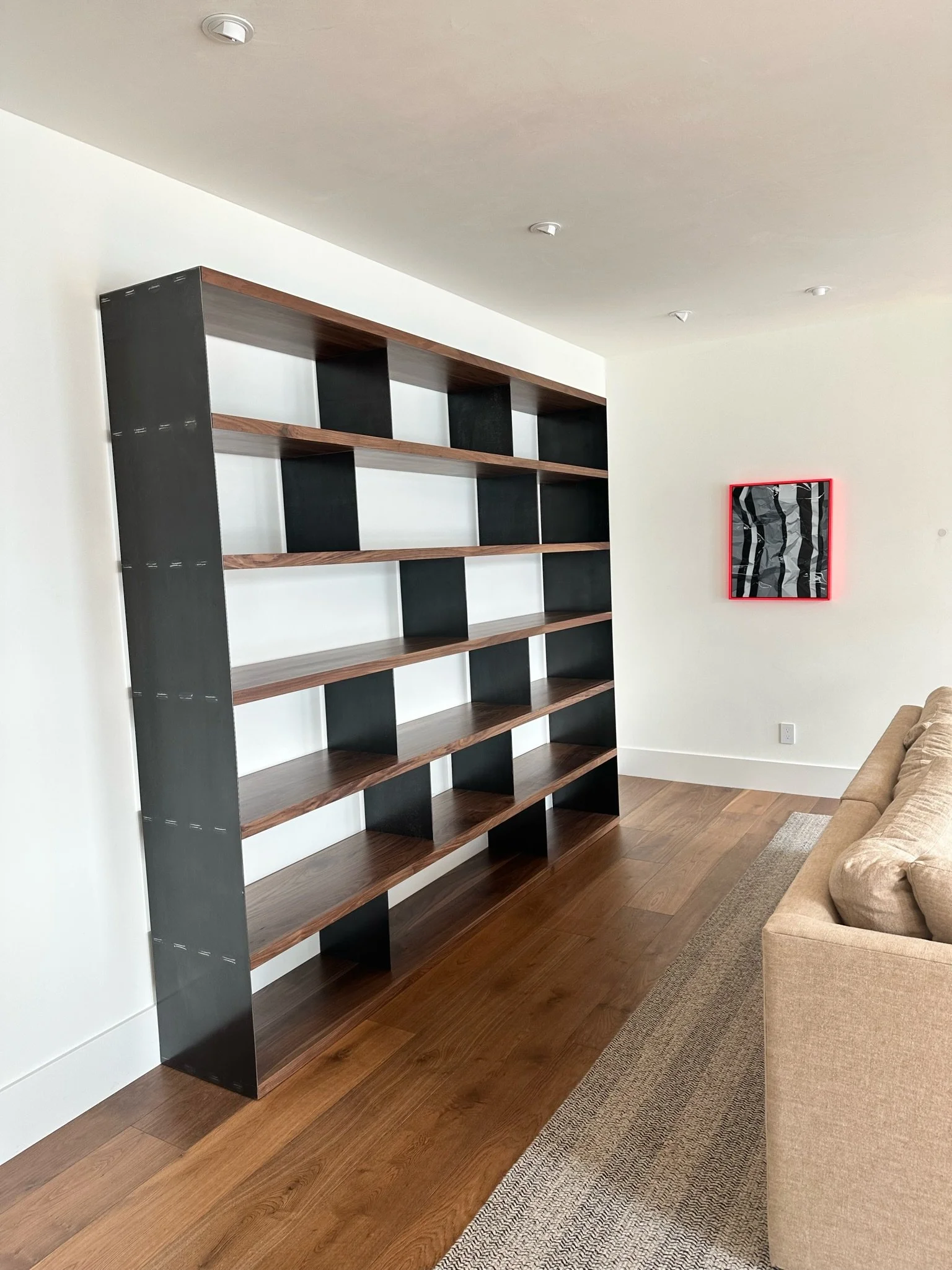 Empty black and wooden bookshelf against a white wall in a living room, with a beige sofa and a patterned rug on wooden floor.