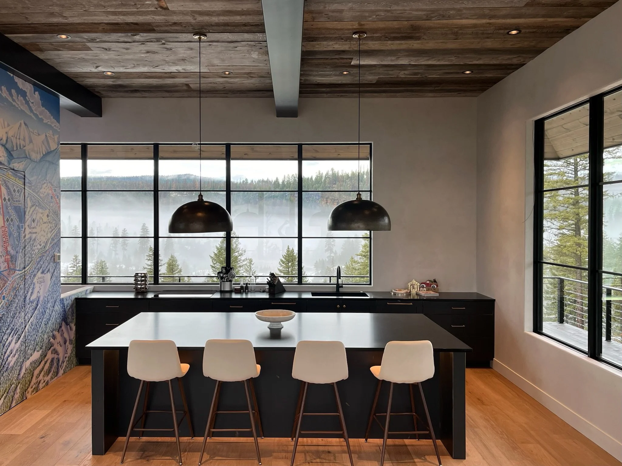 Modern kitchen with large windows overlooking foggy mountains, featuring a black kitchen island, white chairs, pendant lights, wooden ceiling, and a colorful mountain map on the wall.