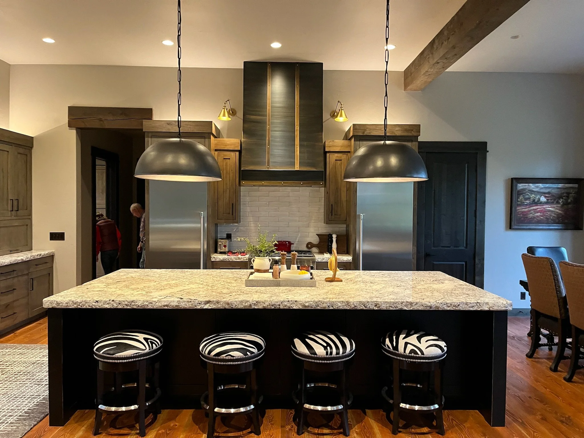 A modern kitchen with a large black island counter with zebra-patterned bar stools, wood cabinets, two gold wall sconces, a stainless steel refrigerator, and a brick backsplash. Two black pendant lights hang over the island, and there are people visi