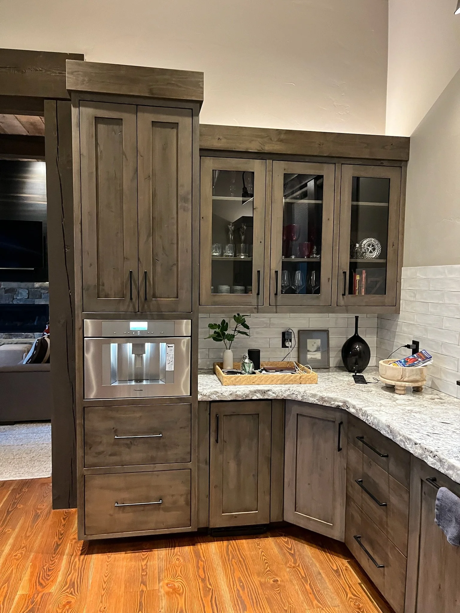 Kitchen with wooden cabinets, granite countertop, and hardwood flooring. Glass cabinet doors display glasses and dishes. Decor includes a small framed picture, black vase, and a basket on the counter.