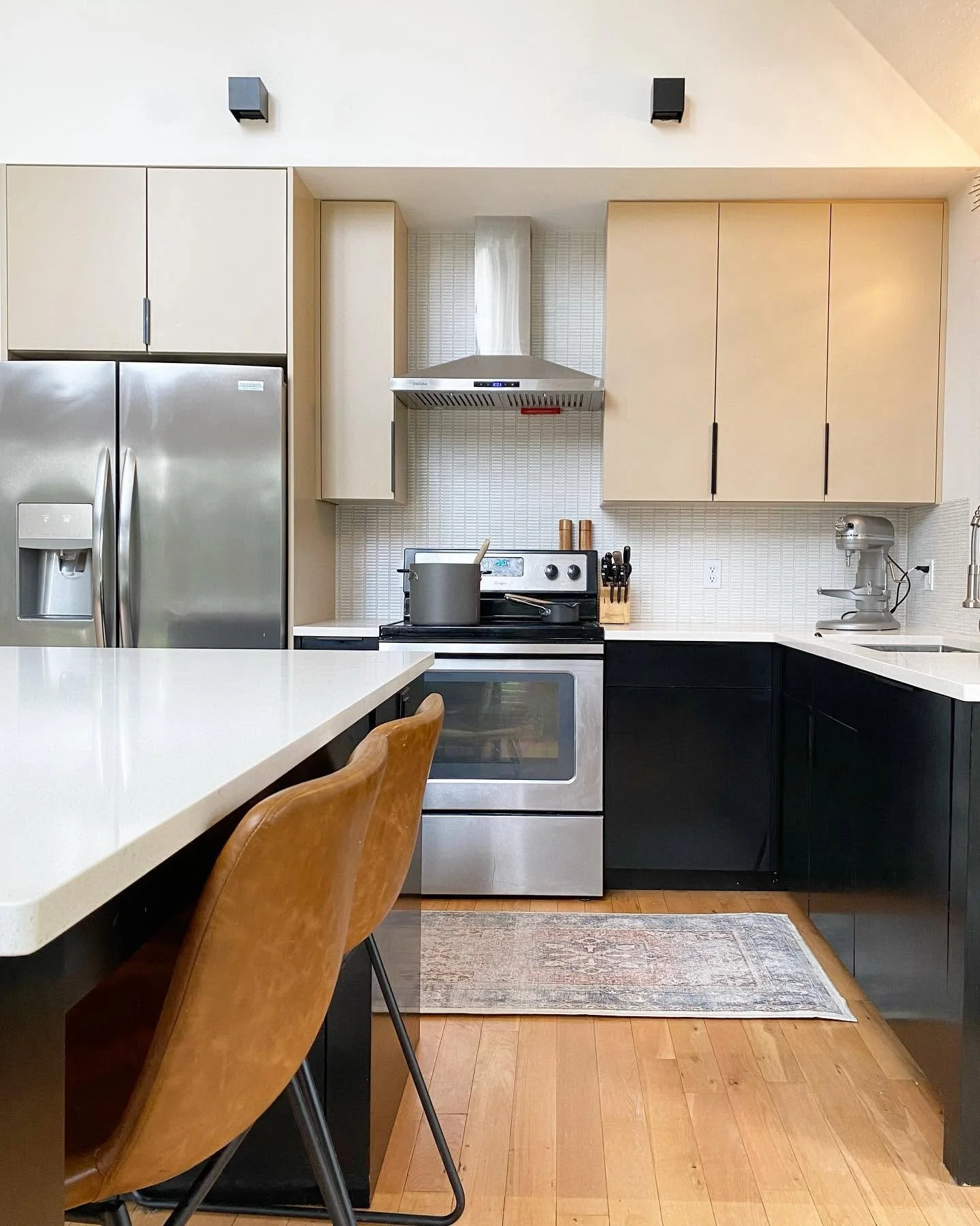Modern kitchen with beige and black cabinets, stainless steel refrigerator, stove with pot, knife set, stand mixer, and white countertop island