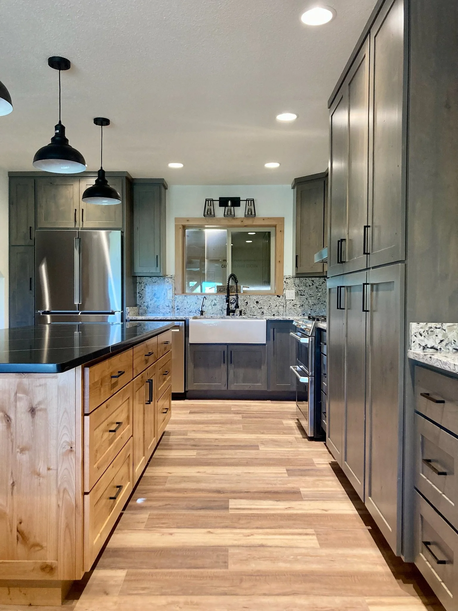 A modern kitchen with wooden and gray cabinets, a granite countertop, a stainless steel refrigerator, black pendant lights, and hardwood flooring.