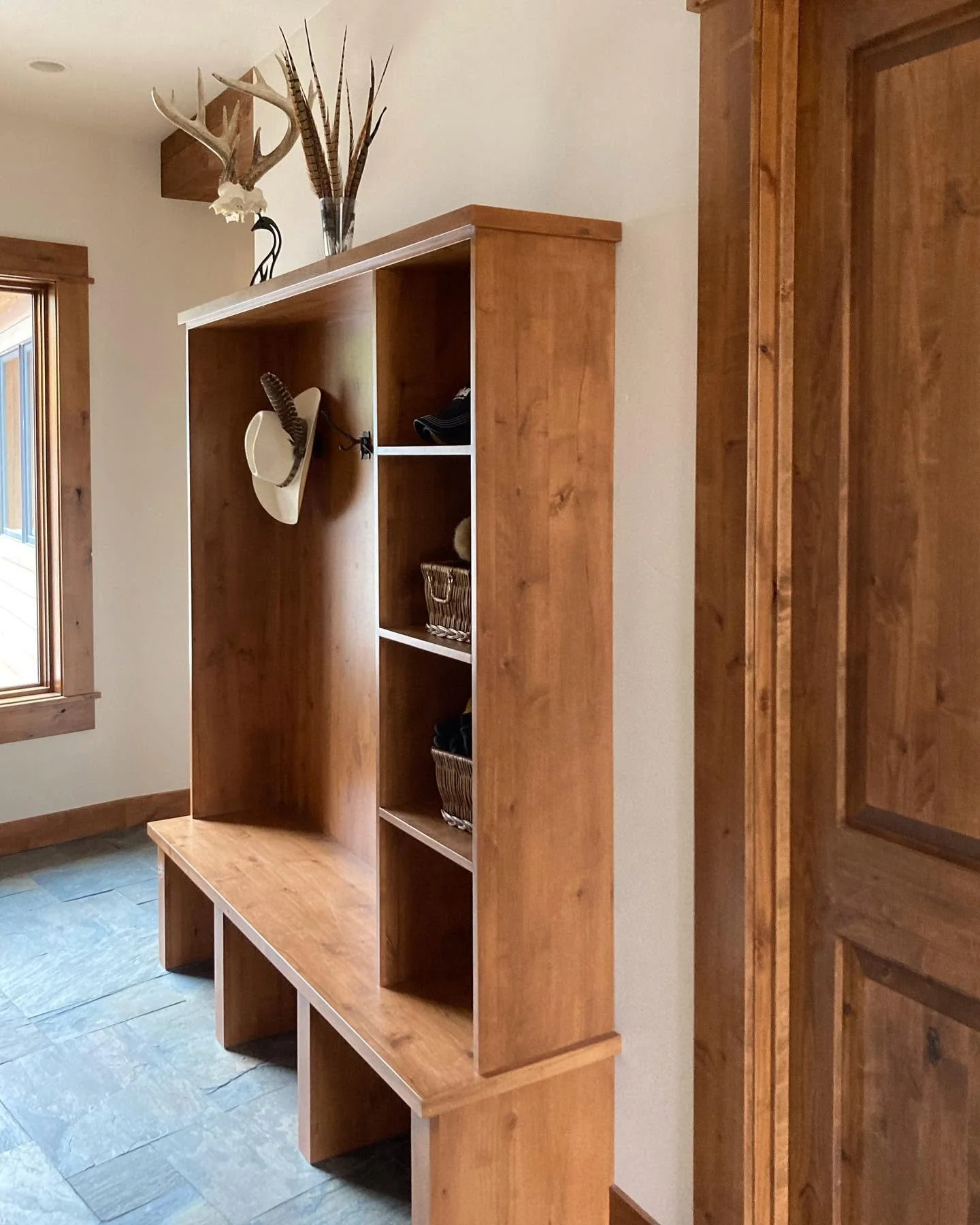 Wooden entryway bench with bookshelves and a decorative antler and owl arrangement on top, next to a window and a wooden door.