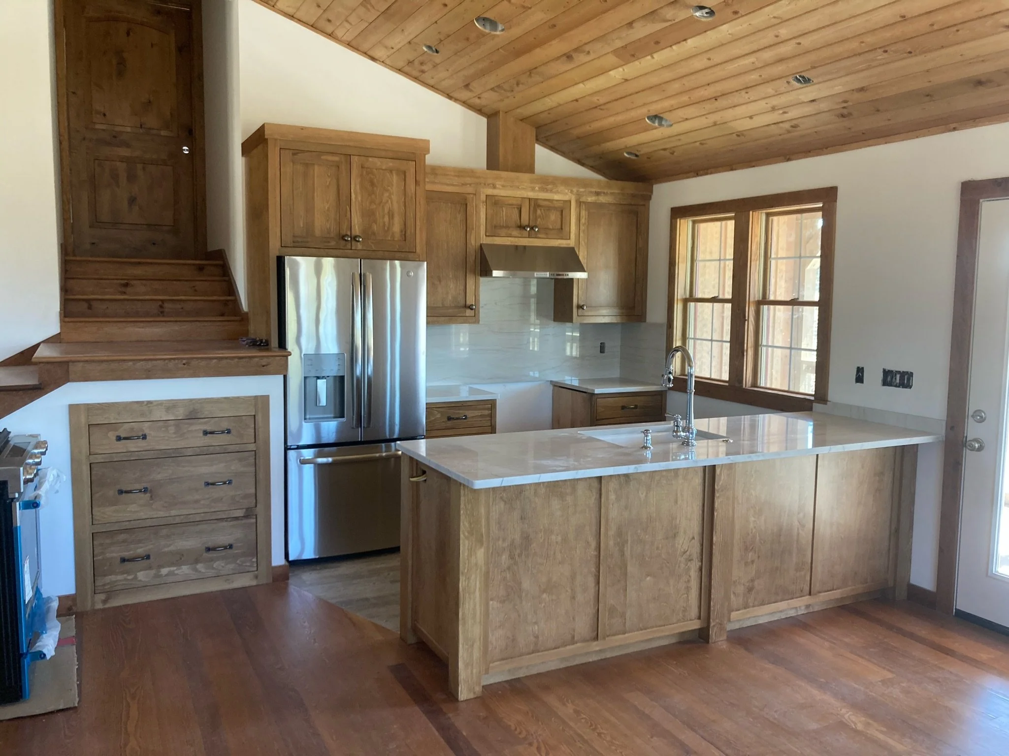Wooden kitchen with a kitchen island, stainless steel refrigerator, upper and lower wooden cabinets, double window, and wooden ceiling.