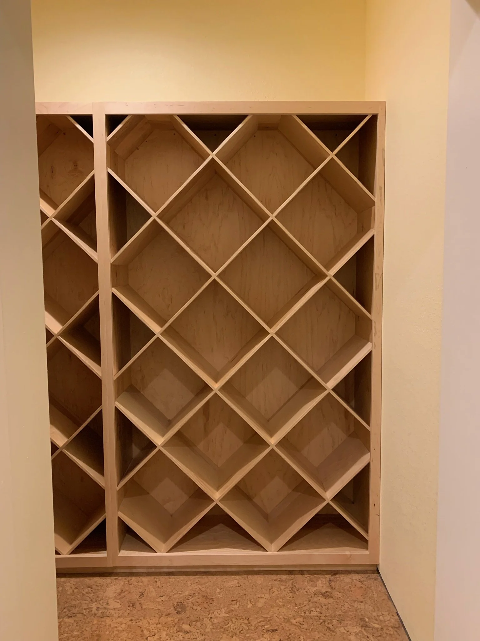 Empty wooden wine rack with diagonal slots, positioned against a yellow wall on a cork floor.
