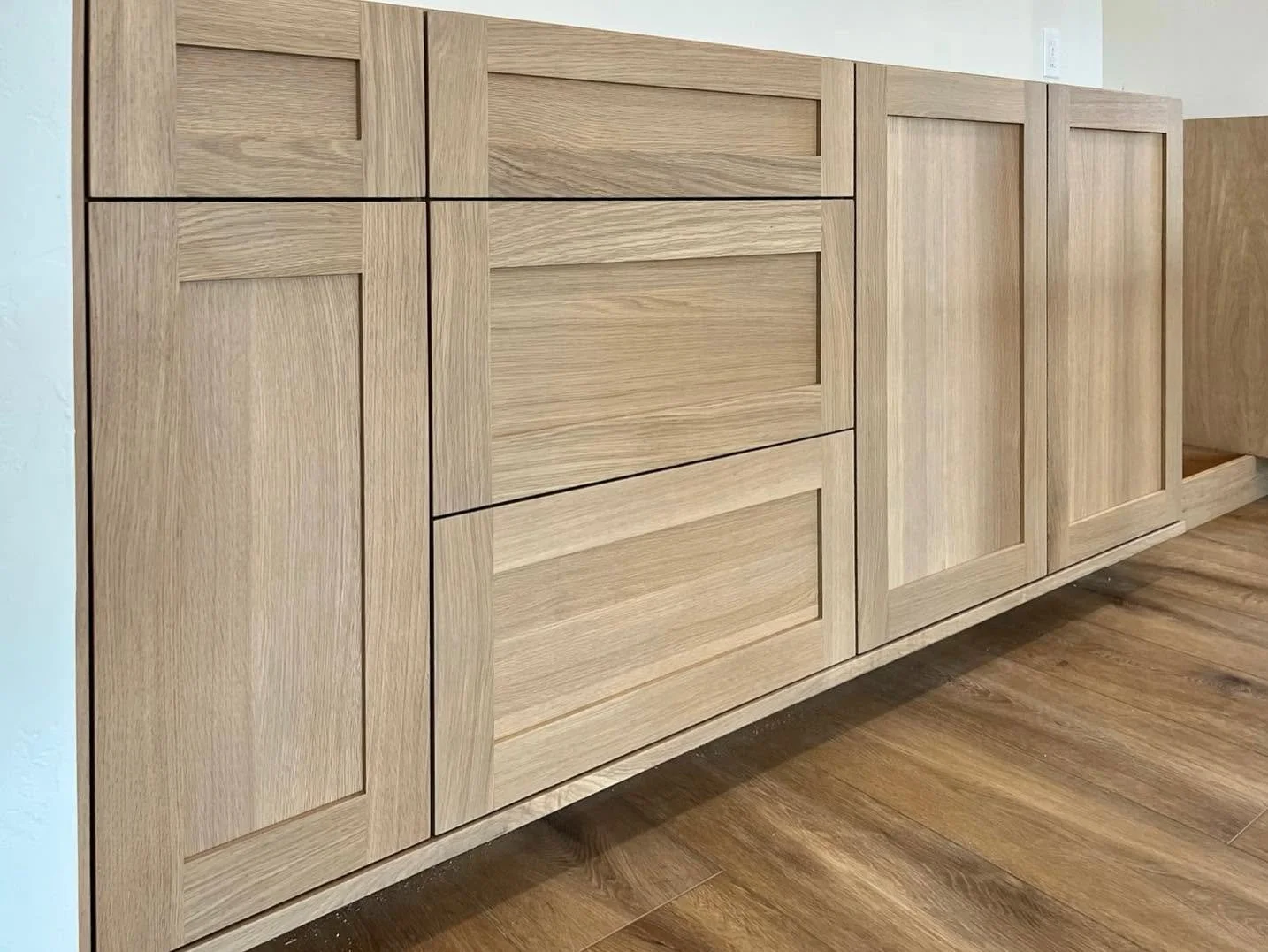 Light wood built-in cabinet with paneled doors and drawers, situated against a white wall with wood flooring.