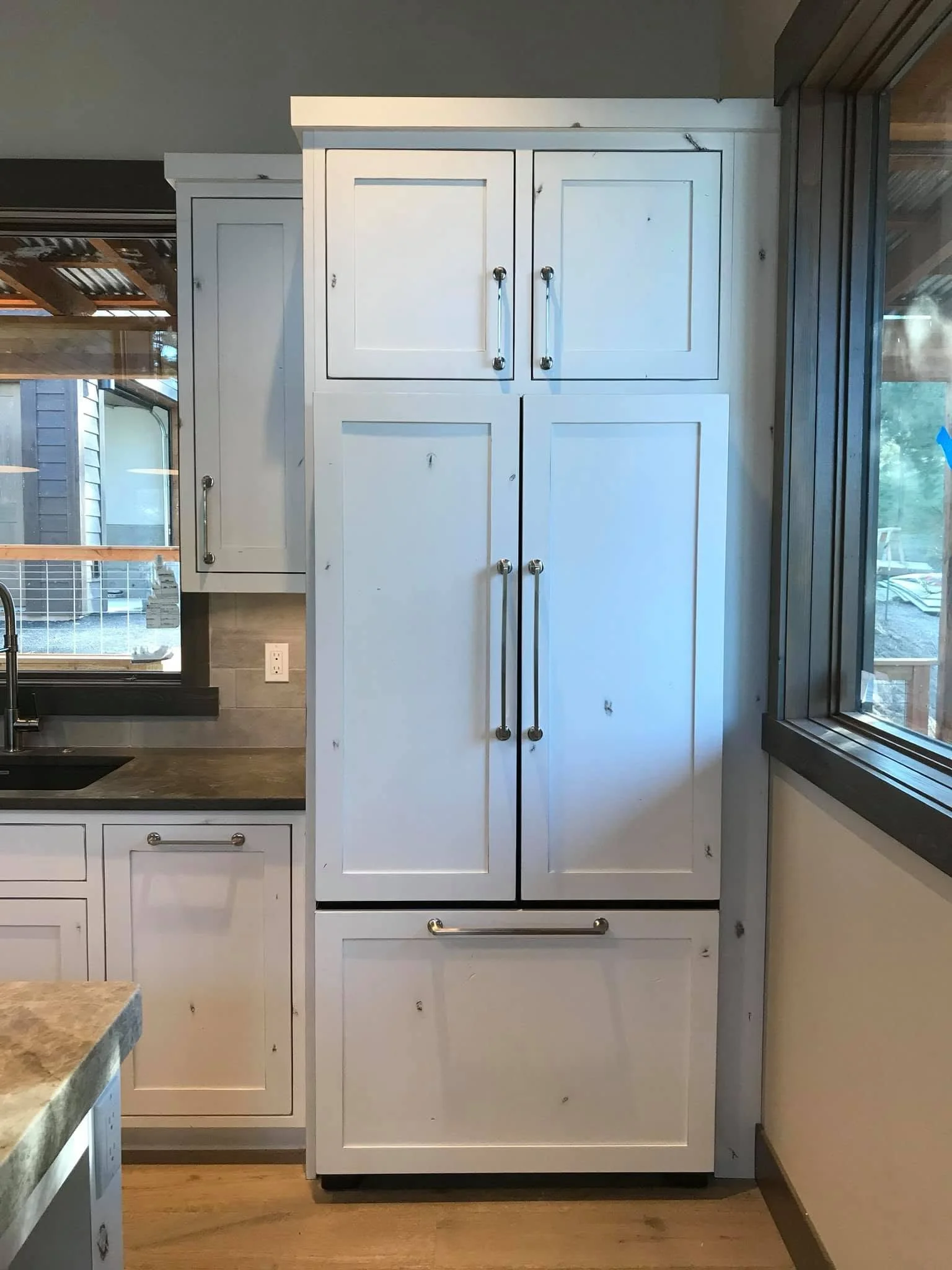 White kitchen cabinet with four doors and silver handles, next to a window with wooden trim, a granite countertop, and a gray tiled wall.