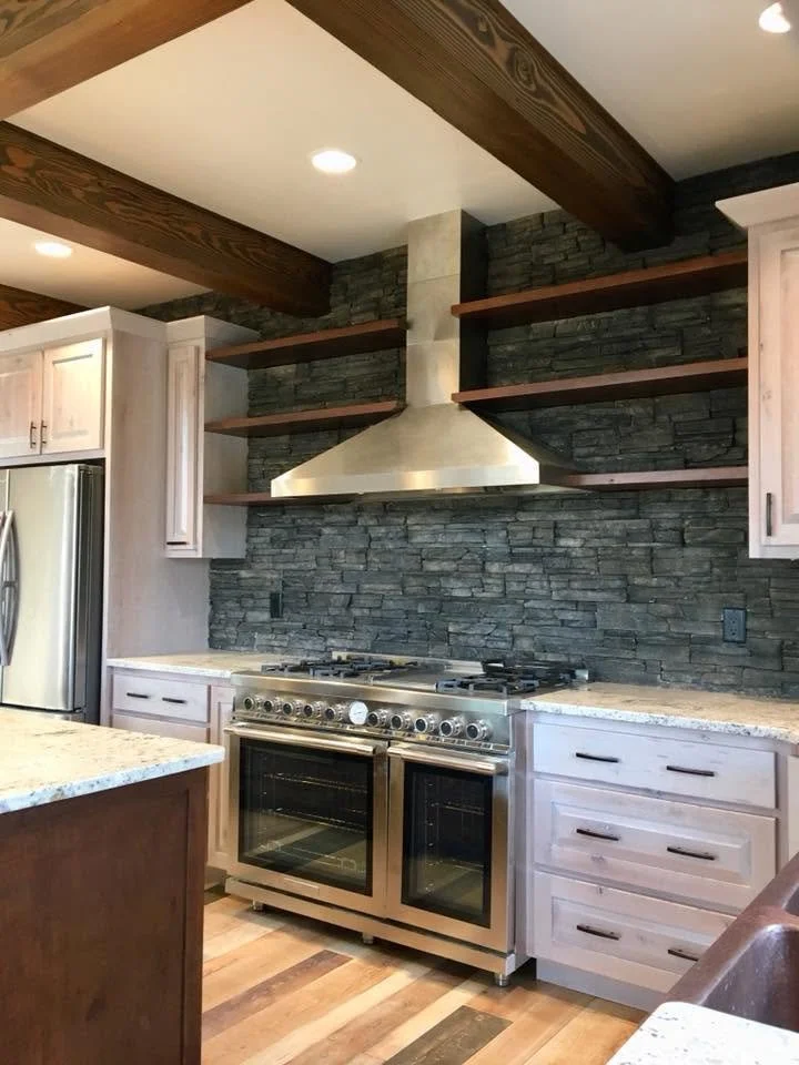 Modern kitchen with stone accent wall, stainless steel range and range hood, open wooden shelves, light-colored cabinetry, and a wooden-beam ceiling.