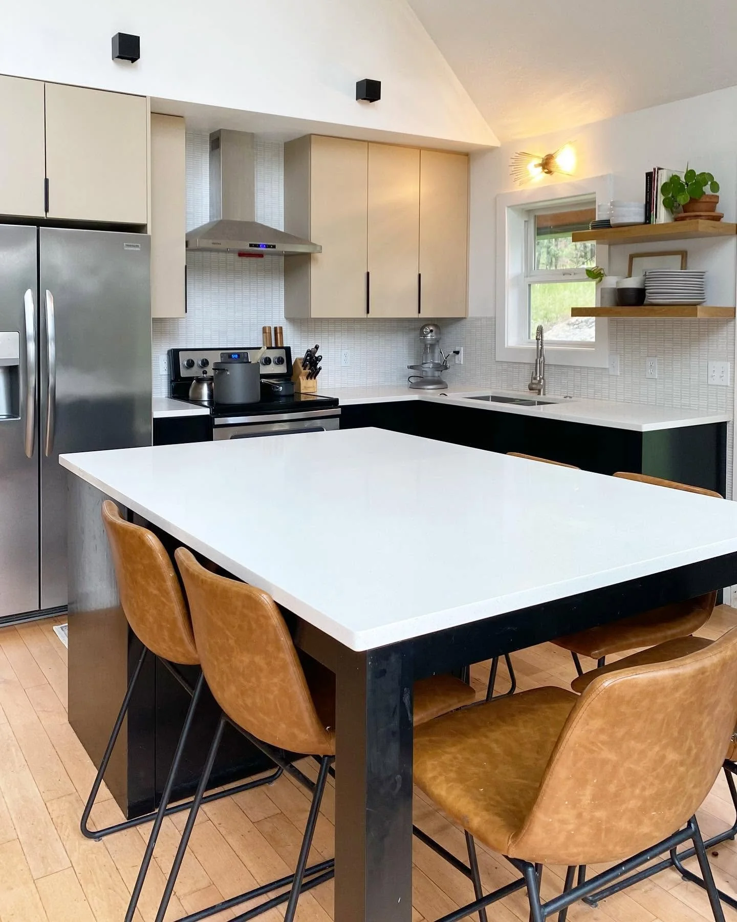 Modern kitchen with a white island countertop, wooden chairs, stainless steel refrigerator, and open wooden shelves with dishes and plants.