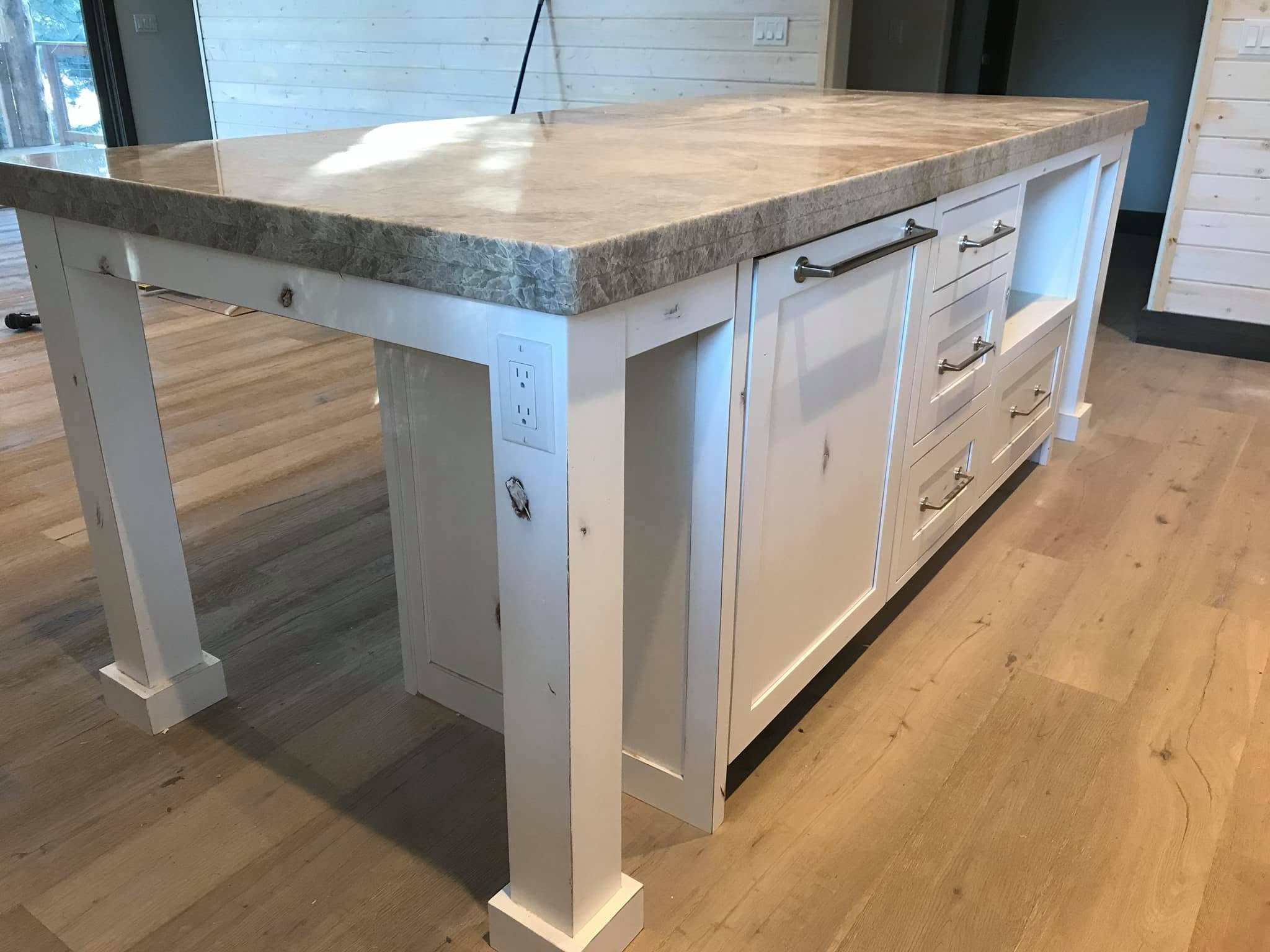 Kitchen island with a marble countertop and white wooden cabinetry, drawers, and electrical outlets, situated in a modern kitchen with wooden flooring and a sliding glass door.