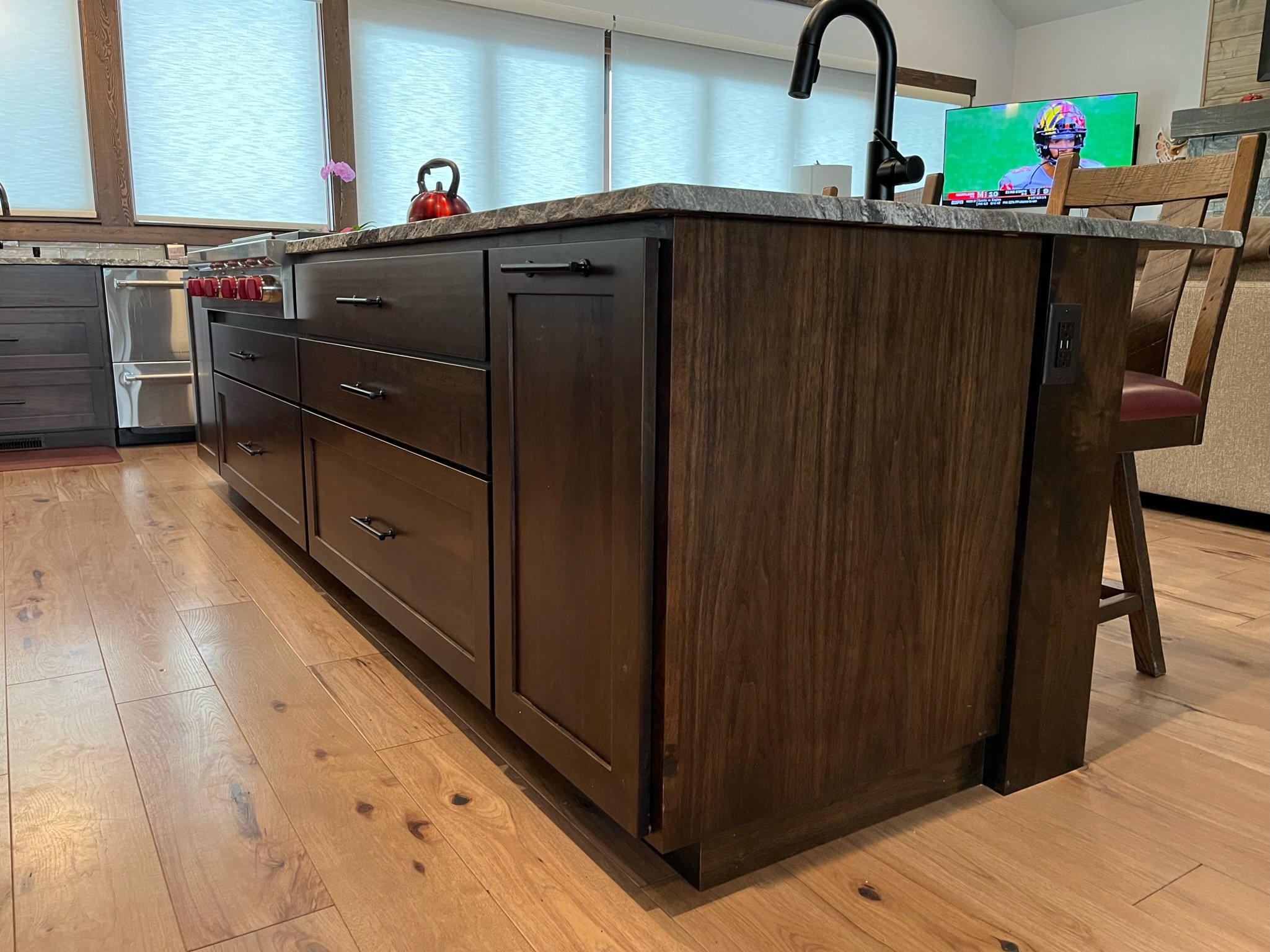 A modern kitchen featuring a large kitchen island with dark wooden cabinets, a granite countertop, a black faucet, and a red kettle on the counter. The background shows a window with frosted glass, a TV displaying a sports broadcast, a wooden dining 