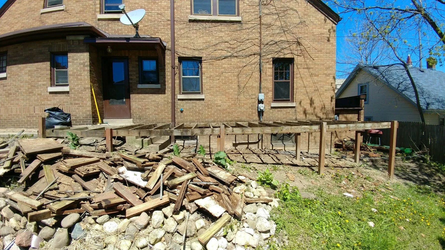 Backyard of a brick house under renovation with wooden deck framing, construction debris, and green plants in the yard. The house has several windows and a satellite dish on the roof.