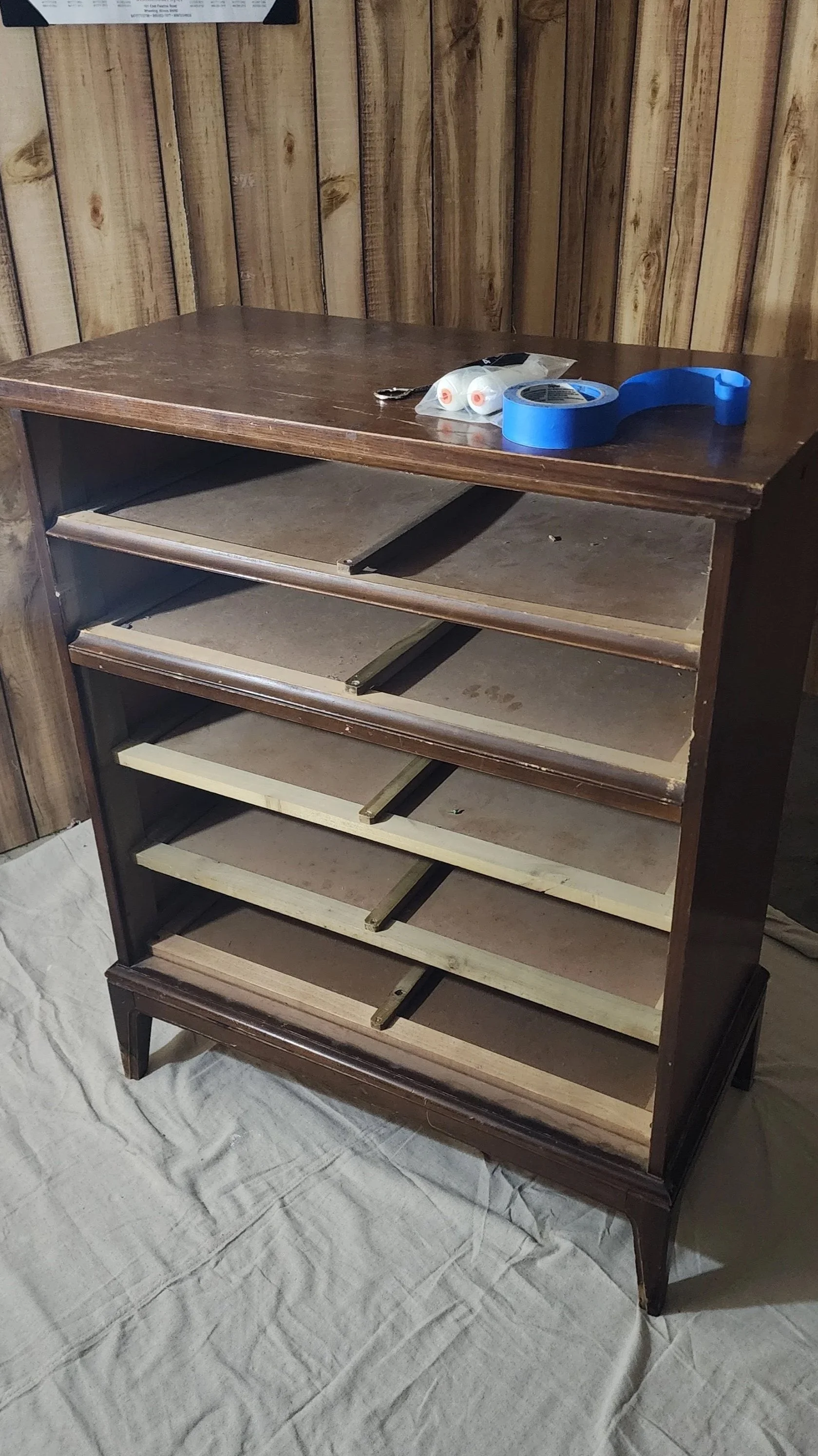 An empty wooden shelf with multiple compartments, on a white cloth, with a blue painter's tape, a small pair of scissors, and two small containers on top, and a wood-paneled wall behind.