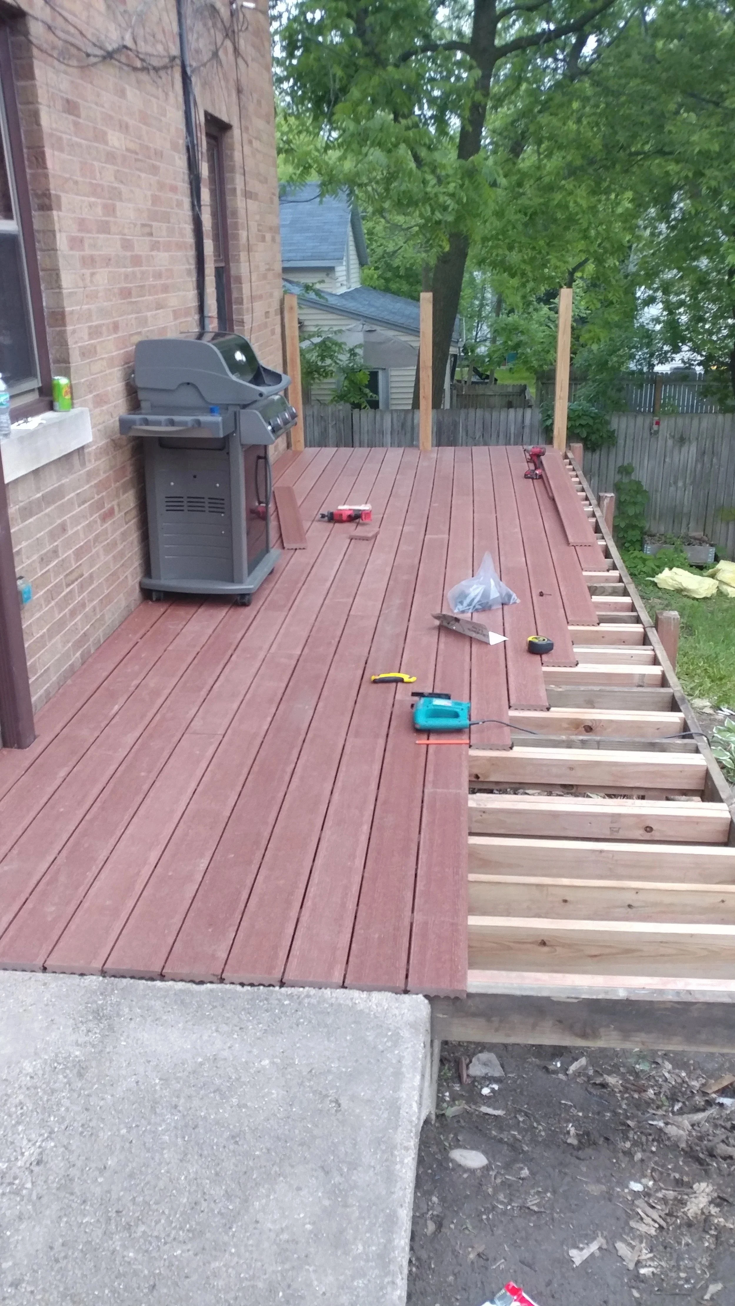 A wooden deck under construction attached to a brick house, with tools and materials scattered on it, including a power drill, screws, and a plastic bag. The deck is partially built, with unfinished framing on one side and a few wooden posts installed around the perimeter.