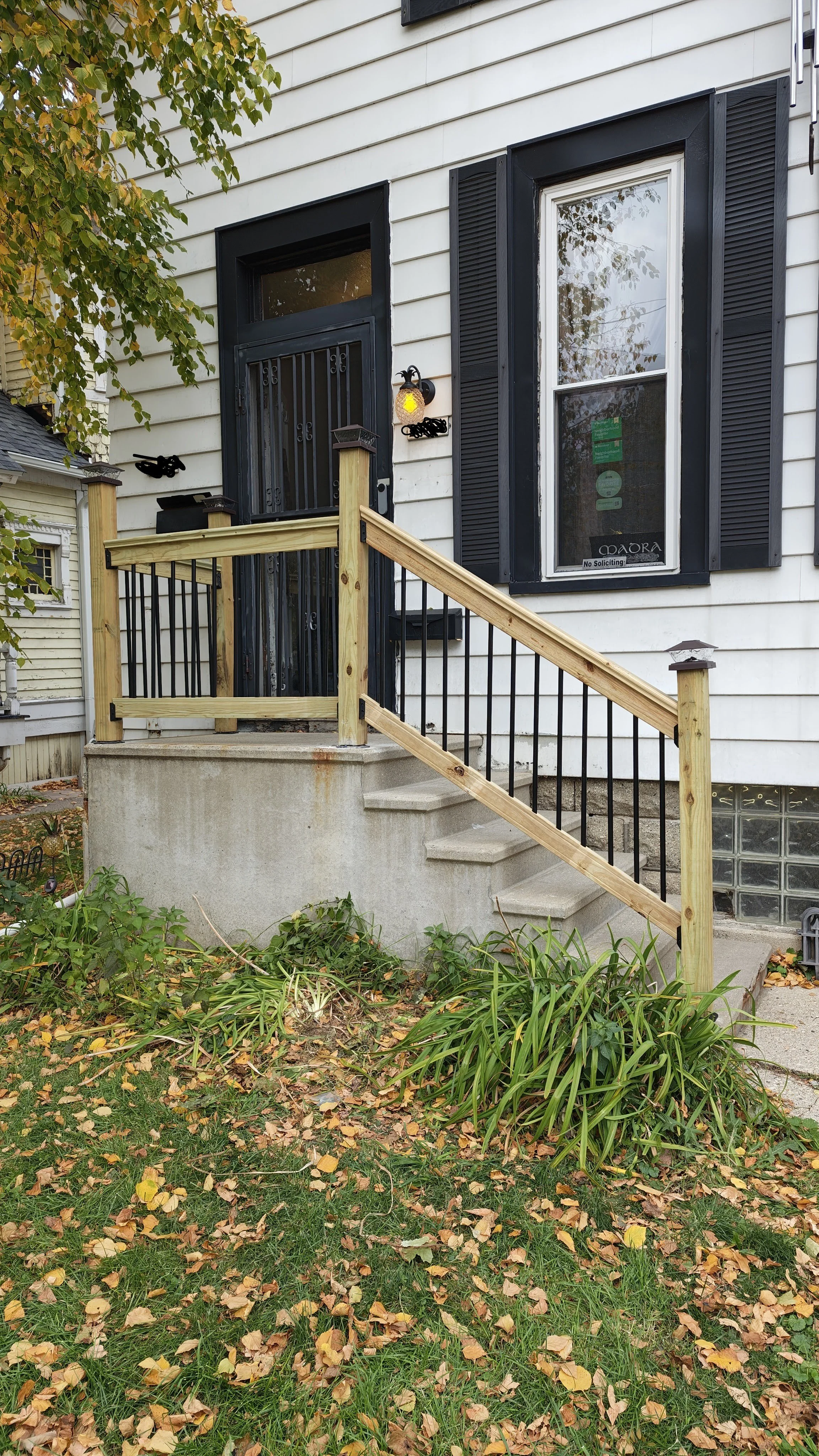 Front porch of a house with a small set of concrete stairs, a newly built wooden railing, a black front door with a security screen, a tall window with black shutters, and some green plants and fallen leaves in the yard.