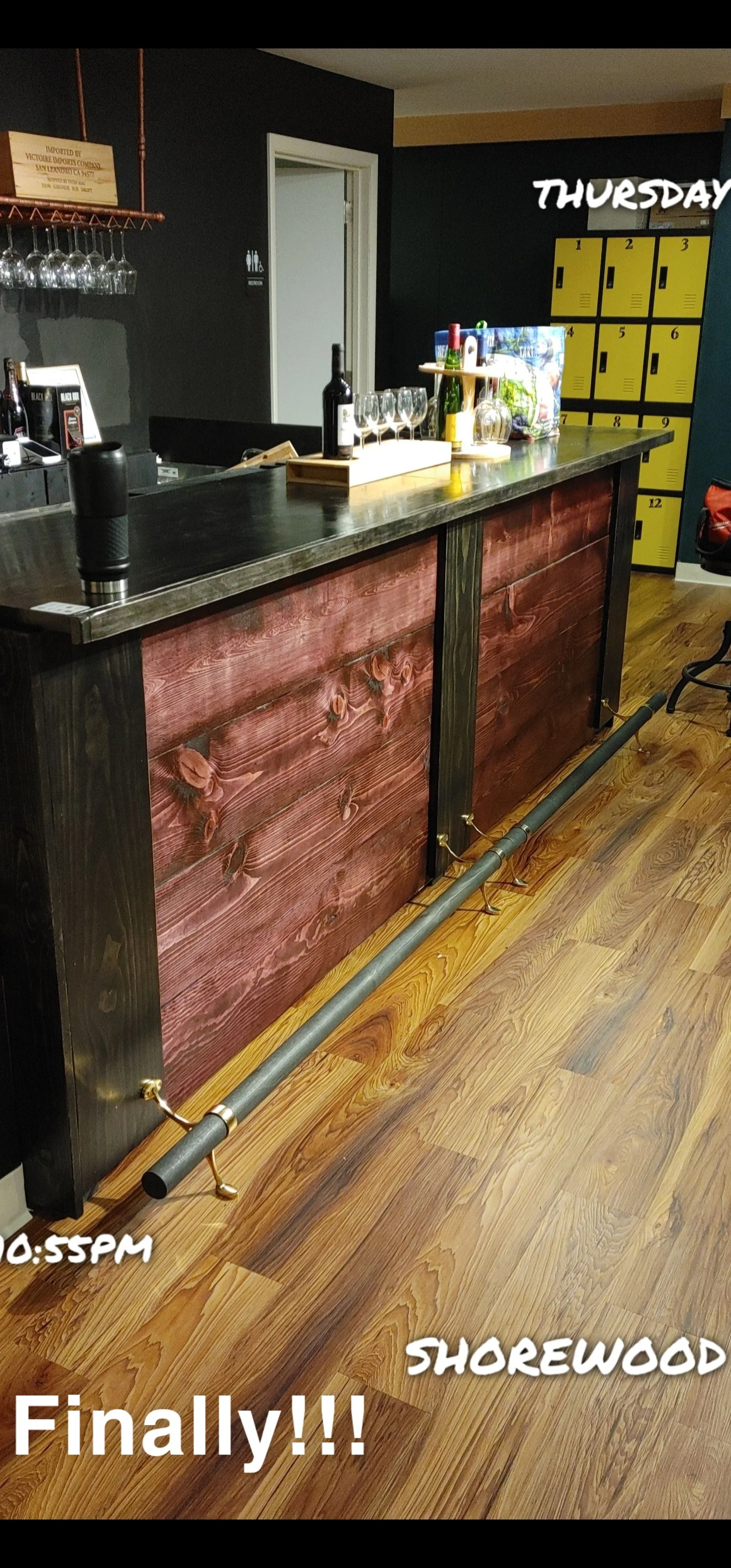 A bar with a wooden front, glasses hanging upside down, bottles of wine, and a row of yellow lockers in the background.