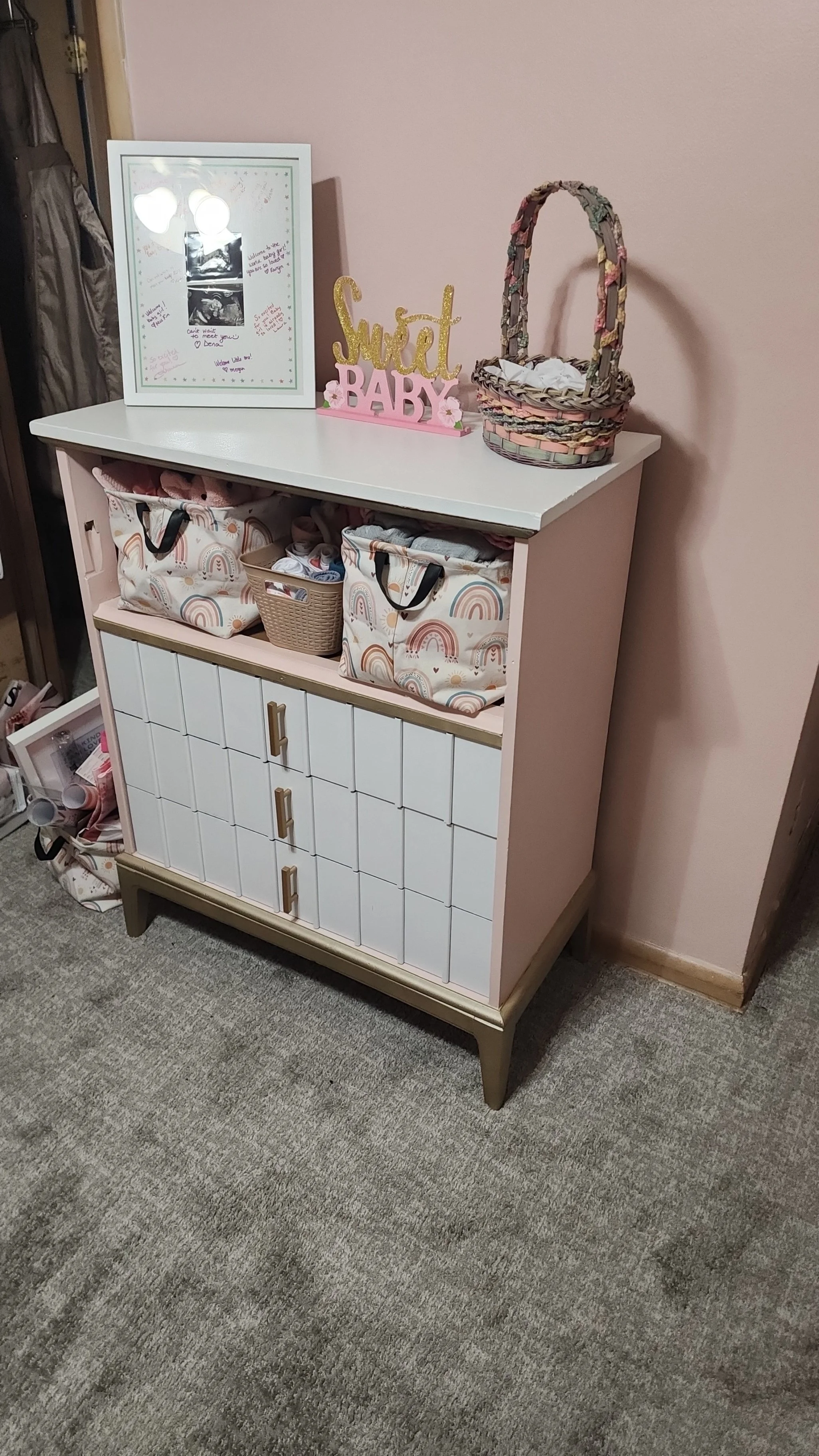 A white and pink dresser with storage compartments and wooden legs. On top, there's a framed ultrasound photo, a pink and gold "Sweet Baby" sign, and a decorative basket. Underneath the dresser, there are storage baskets with baby clothes and accessories. The dresser is against a light pink wall with a gray carpeted floor.
