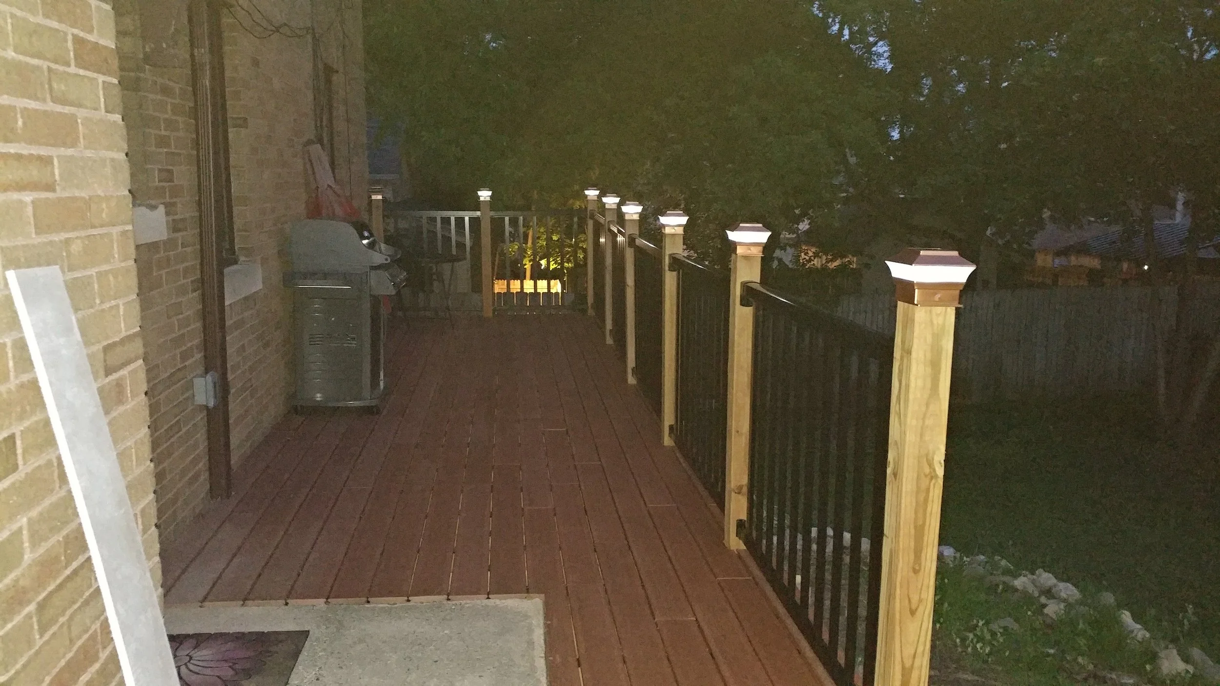A backyard porch at night with a brick wall to the left, a wooden railing with black metal balusters, and a wooded area in the background illuminated by outdoor lighting.