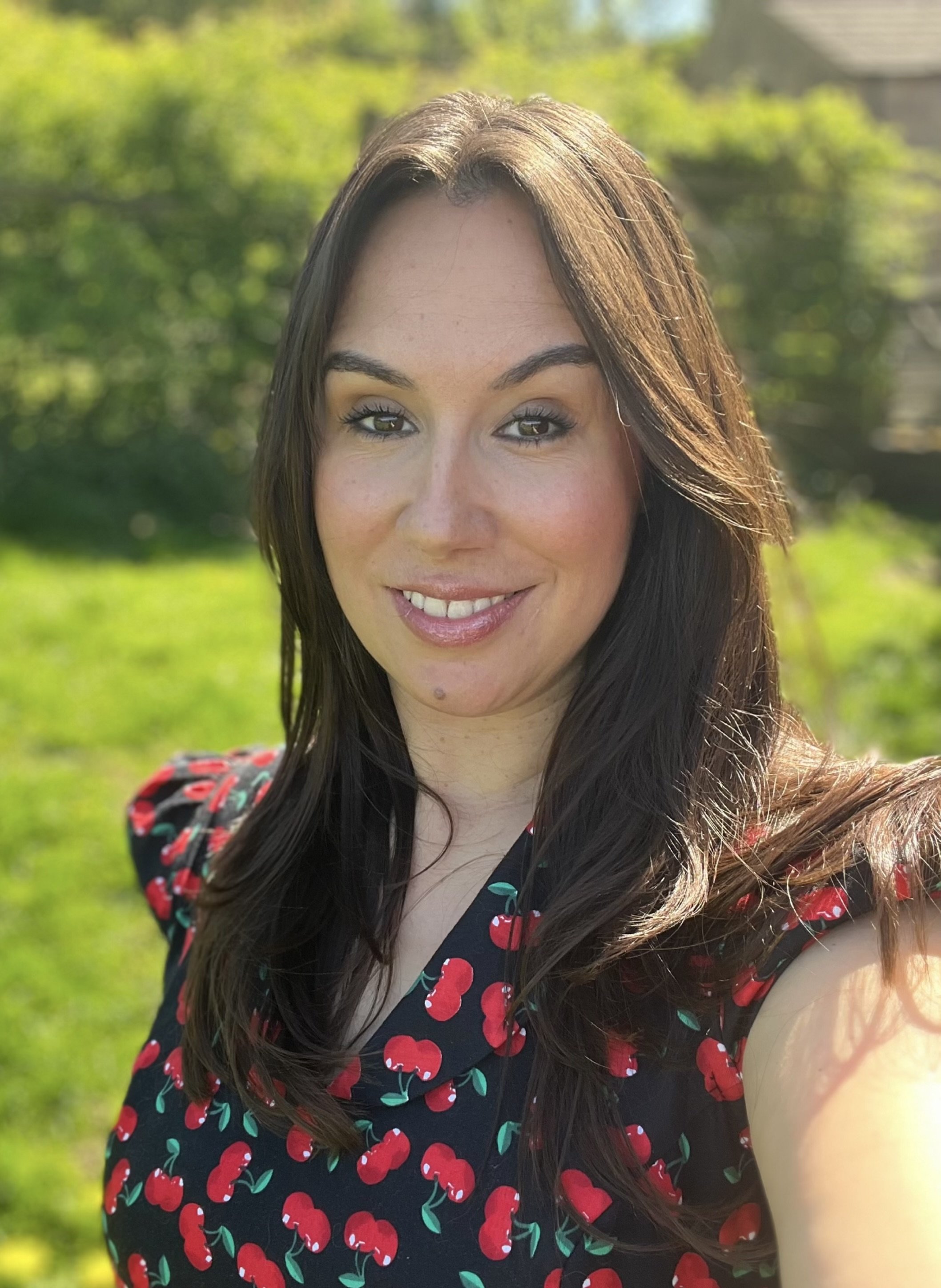 A woman with long dark brown hair, light skin, and brown eyes is smiling at the camera. She is outdoors with a background of greenery and a house, sunny daylight, wearing a black dress with a red cherry pattern.