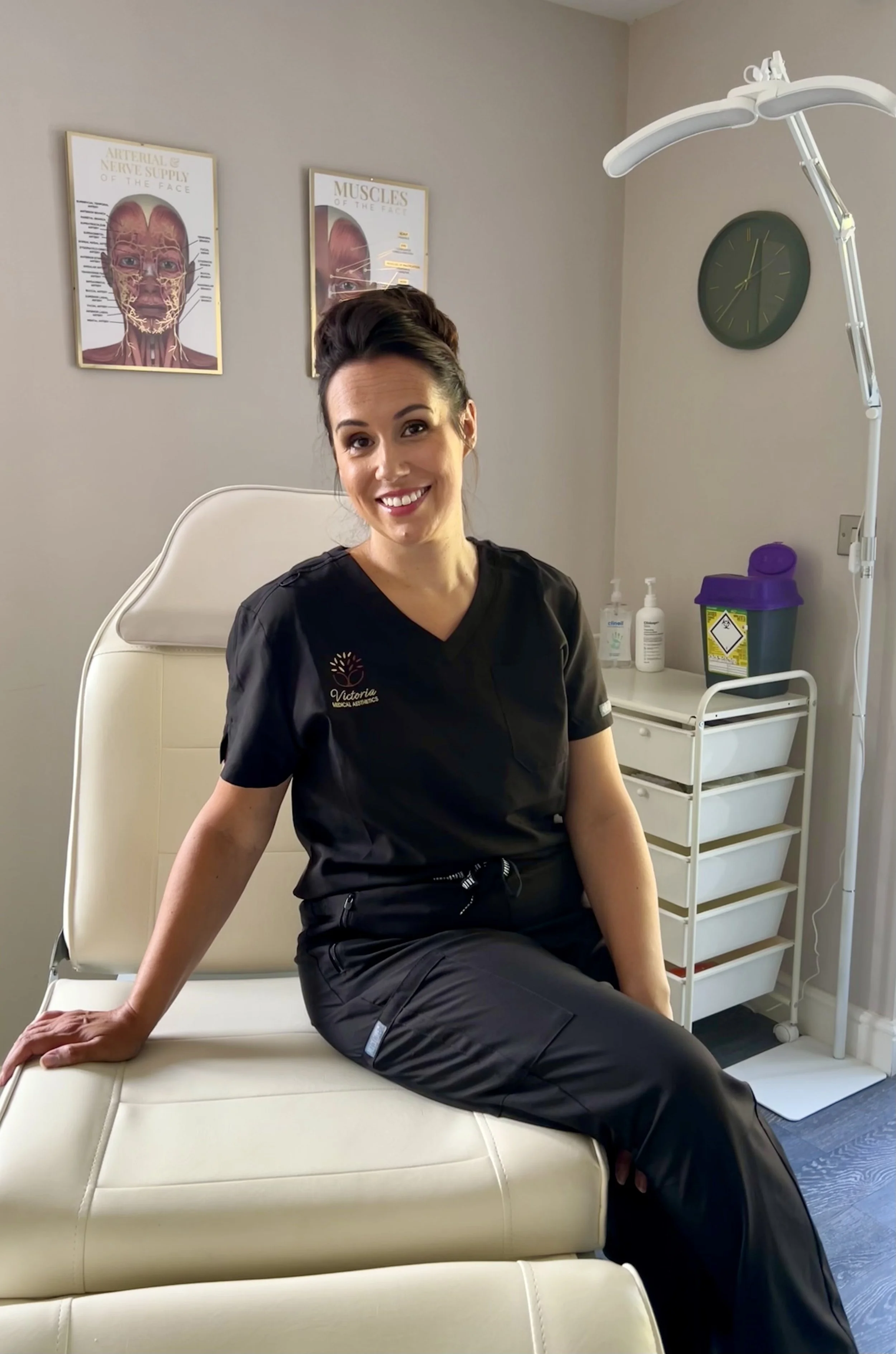 A woman wearing medical scrubs sitting on an examination table in a clinic room, smiling at the camera. Behind her, medical posters about facial anatomy, a wall clock, and medical supplies on a rolling cart are visible.