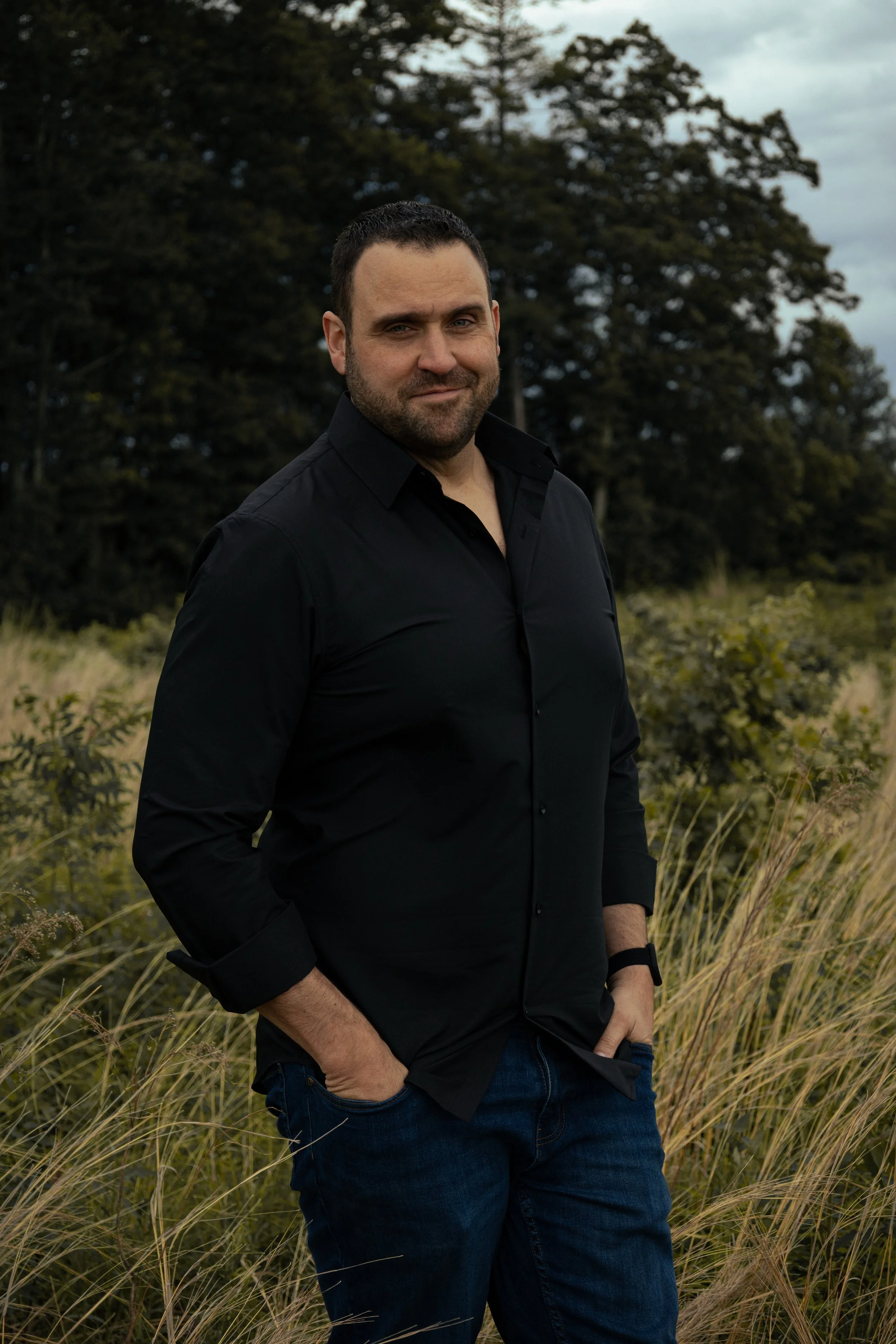 A man with short dark hair and a beard standing outdoors among tall grasses, wearing a black button-up shirt and jeans, with a background of trees and a cloudy sky.
