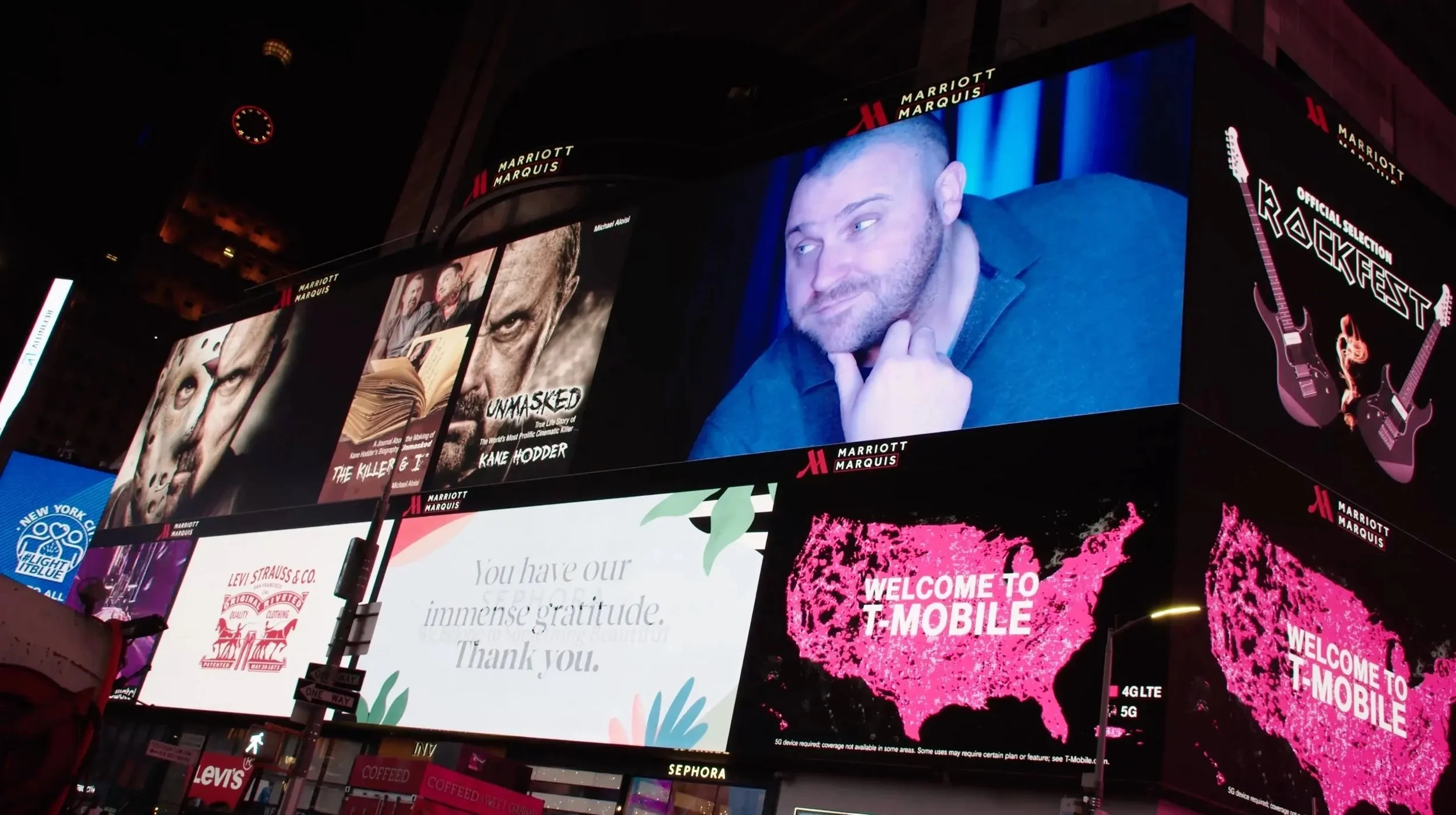 Nighttime photo of a large digital billboard in Times Square displaying various advertisements, including a close-up image of a man with a beard and a shaved head, a poster for the film 'Unmasked' by Michael Aloni, and ads for Marriott Marquis hotel and T-Mobile. The billboard features colorful graphics and text.