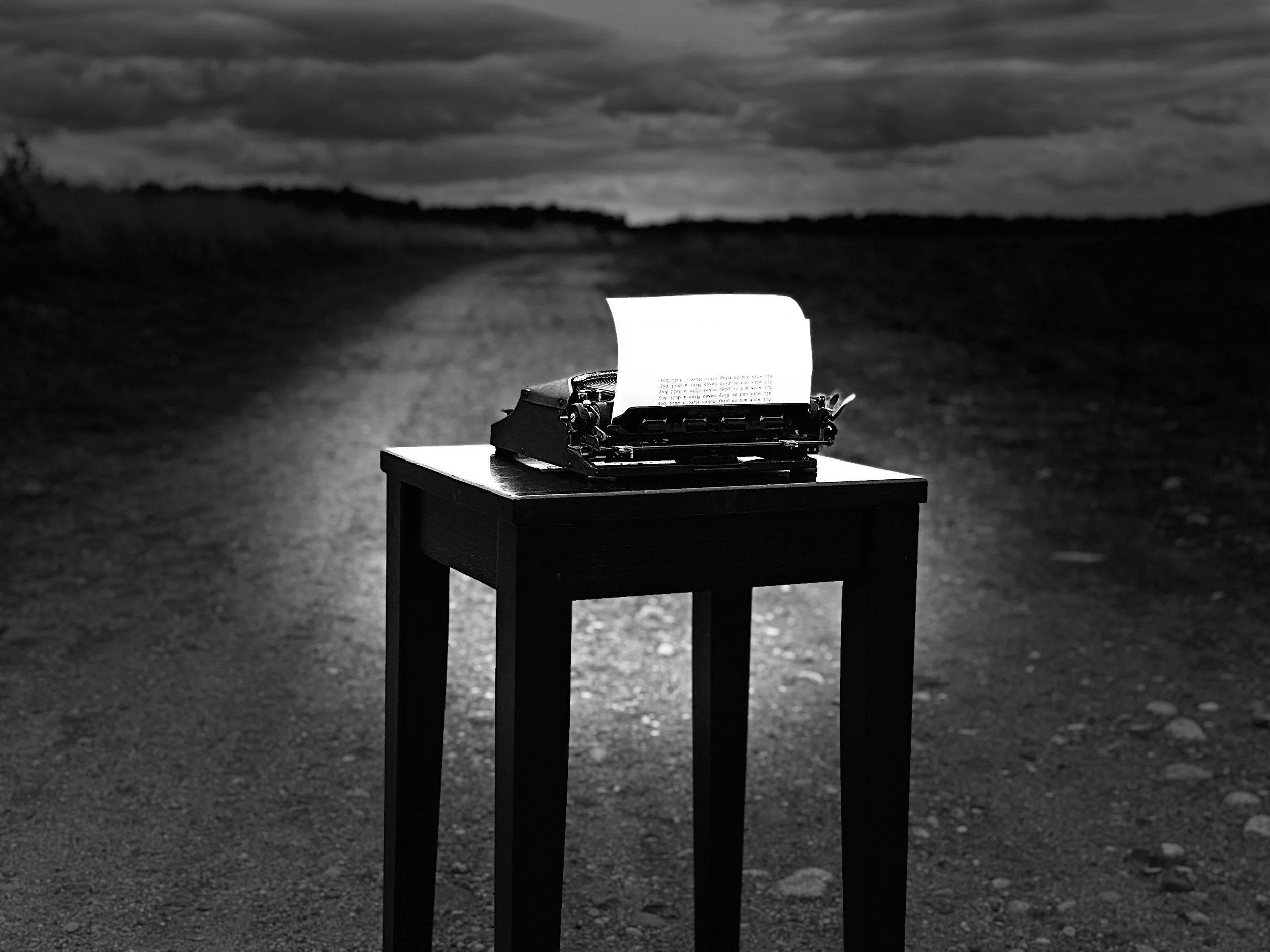 A vintage typewriter on a small table outdoors at dusk or night, with a piece of paper inserted, set against a dark, cloudy sky with distant hills or trees.