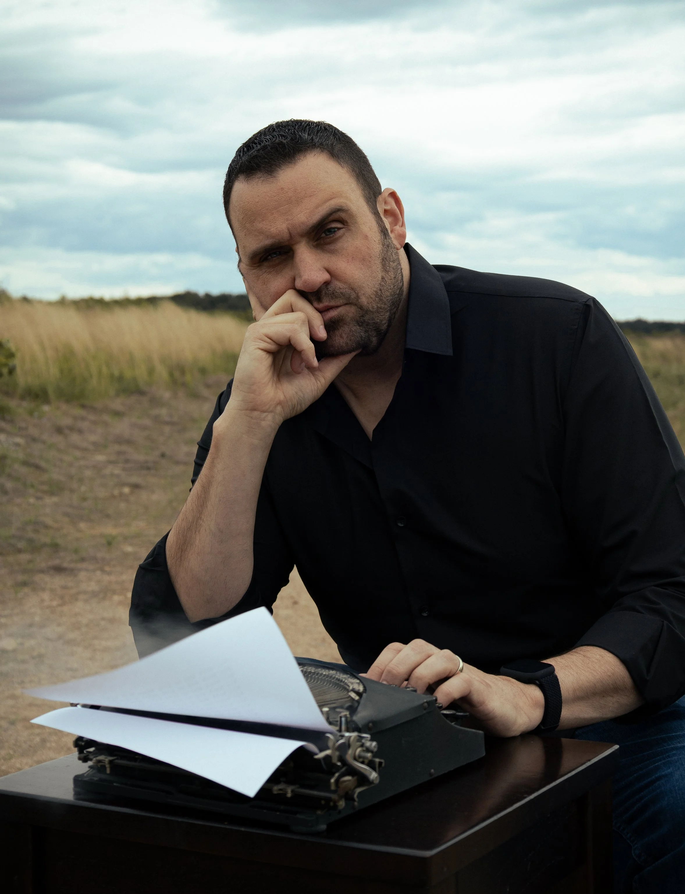 Man with dark hair and beard working on a typewriter outdoors, holding his hand to his face, with papers and a desk in front of him against a cloudy sky background.