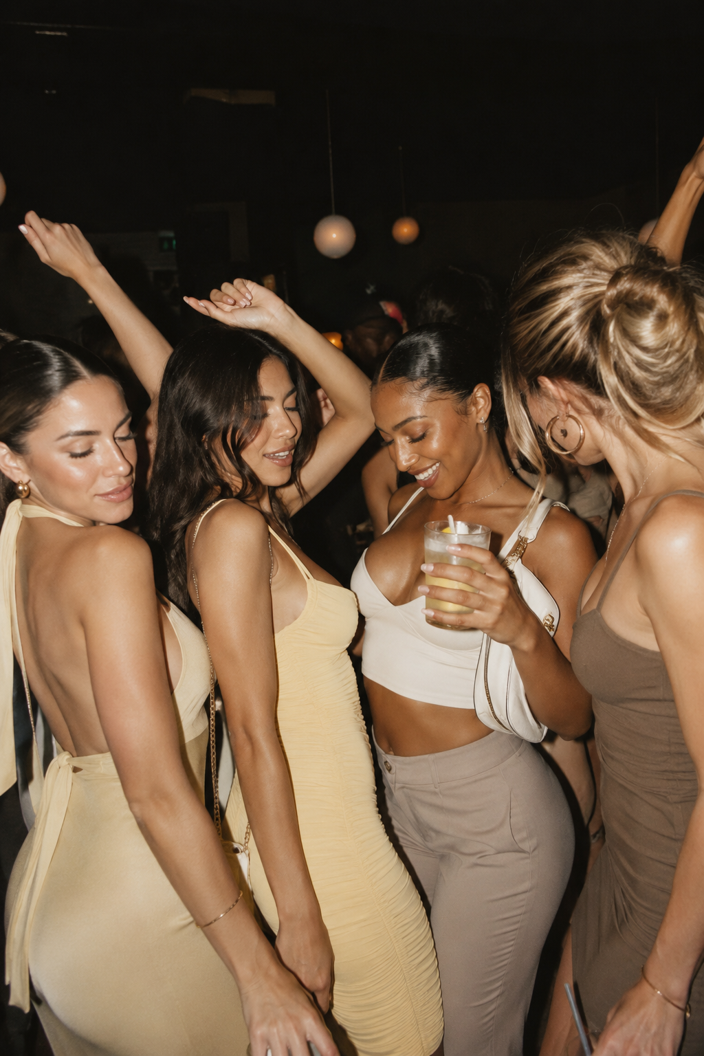 Group of women dancing and socializing at a party or nightclub, wearing stylish outfits and holding drinks.