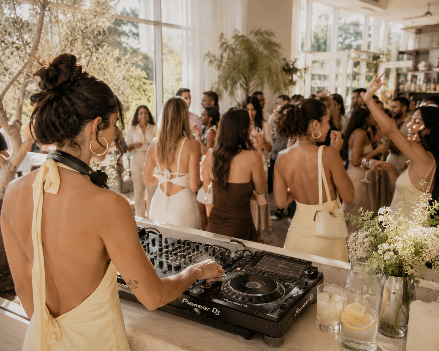 A female DJ with dark hair in an updo and hoop earrings, wearing a backless dress with a bow, plays music at a lively indoor party with many standing people, some holding drinks, in a bright, decorated space with large windows and greenery.