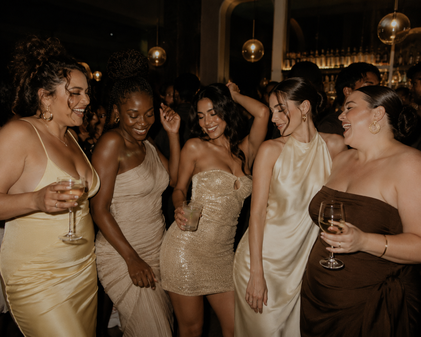 Group of women dancing and having fun at a party, holding glasses of wine or drinks, dressed in elegant evening gowns in a dimly lit venue.