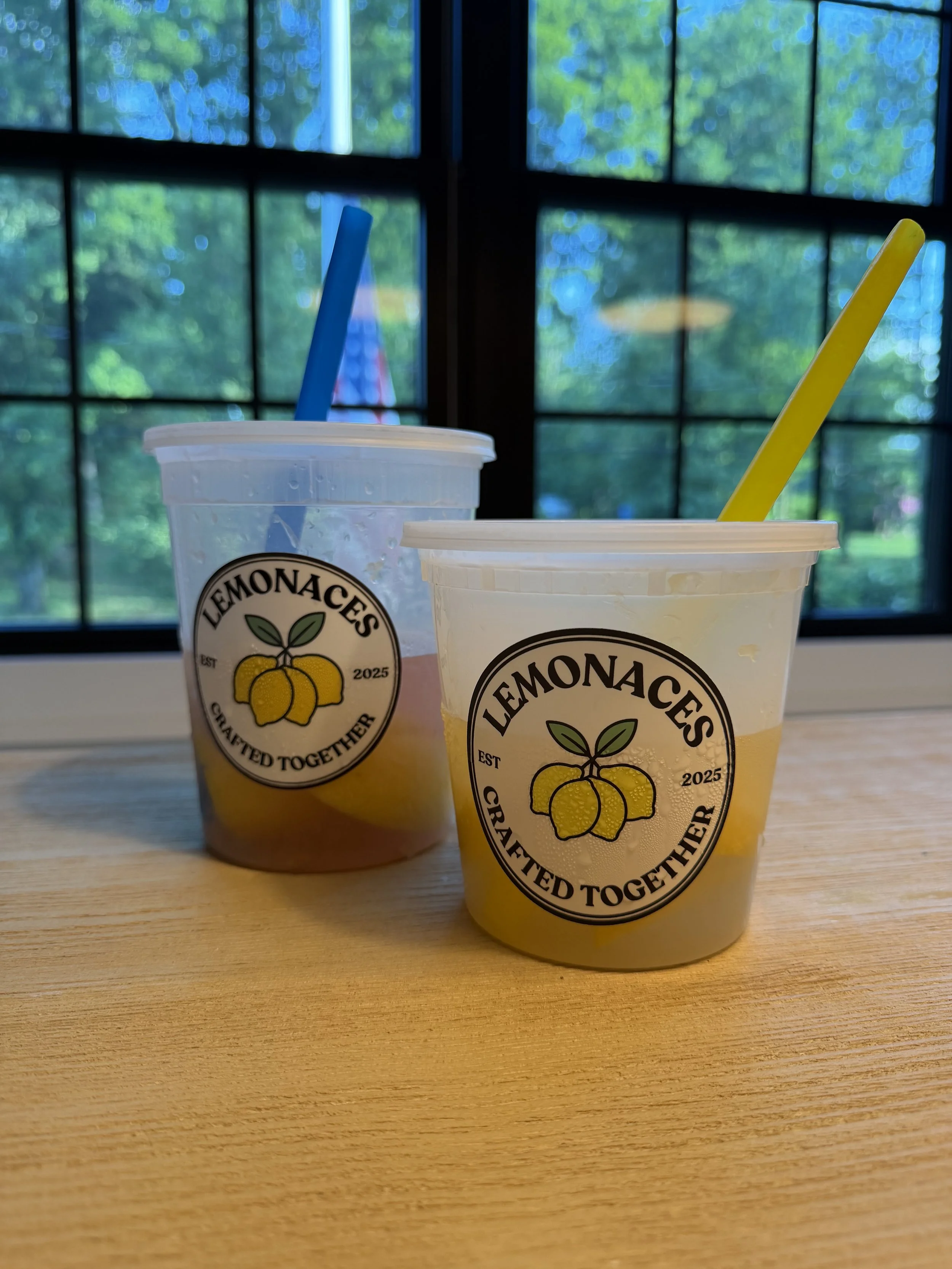 Two cups from LemonaChes, a lemon-themed beverage brand, on a wooden table by a window with a green outdoor scene, one cup with blue straw and the other with yellow straw.