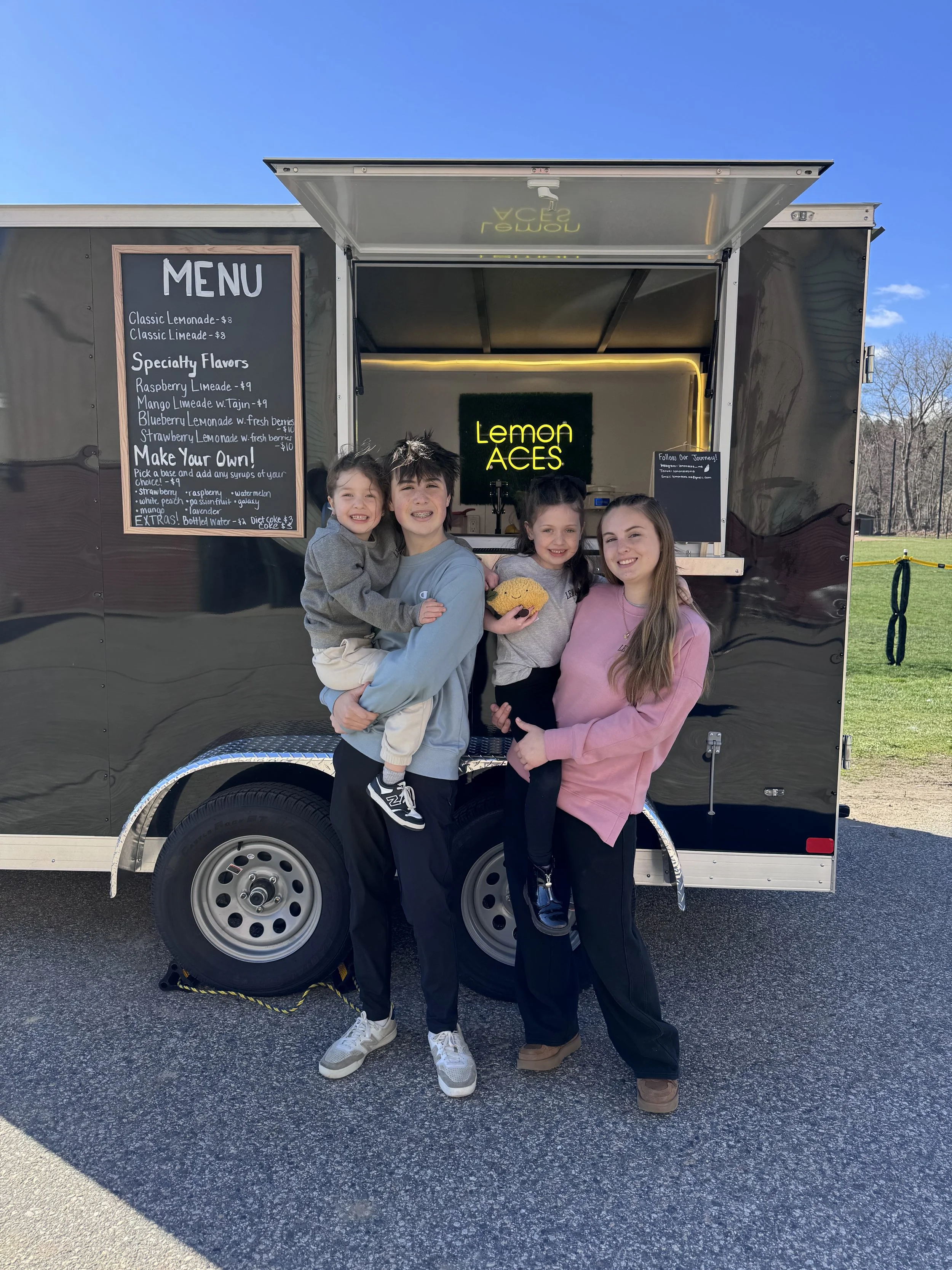 Four people standing in front of a black lemonade stand trailer with a menu board, smiling and posing for the photo outdoors on a sunny day.