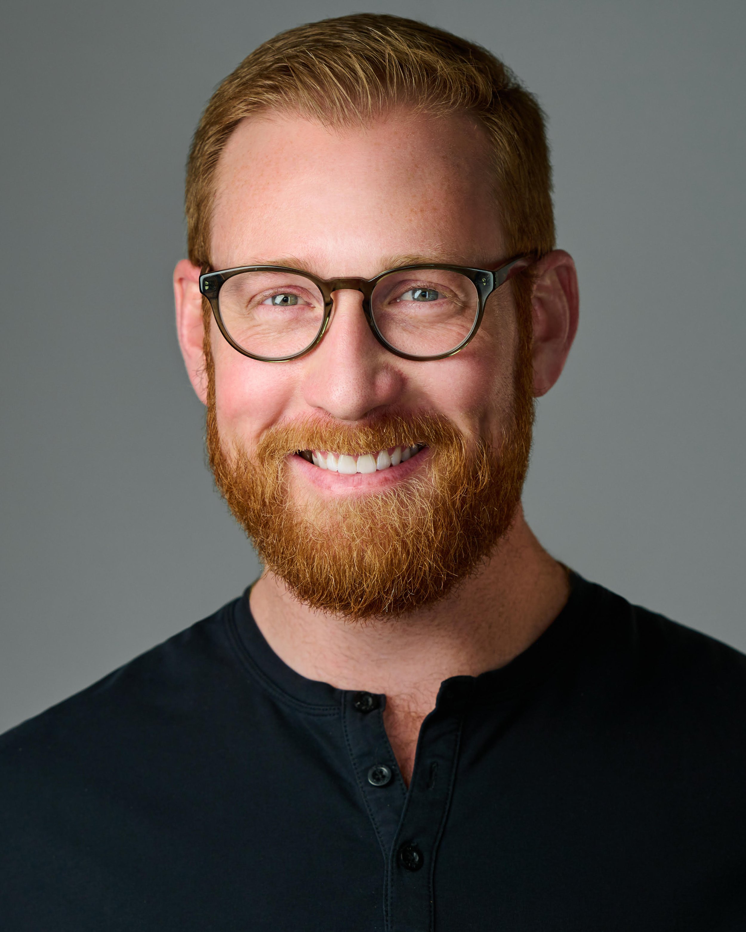 Close-up of a man with glasses, red hair, and a beard, smiling, wearing a black shirt, against a gray background.