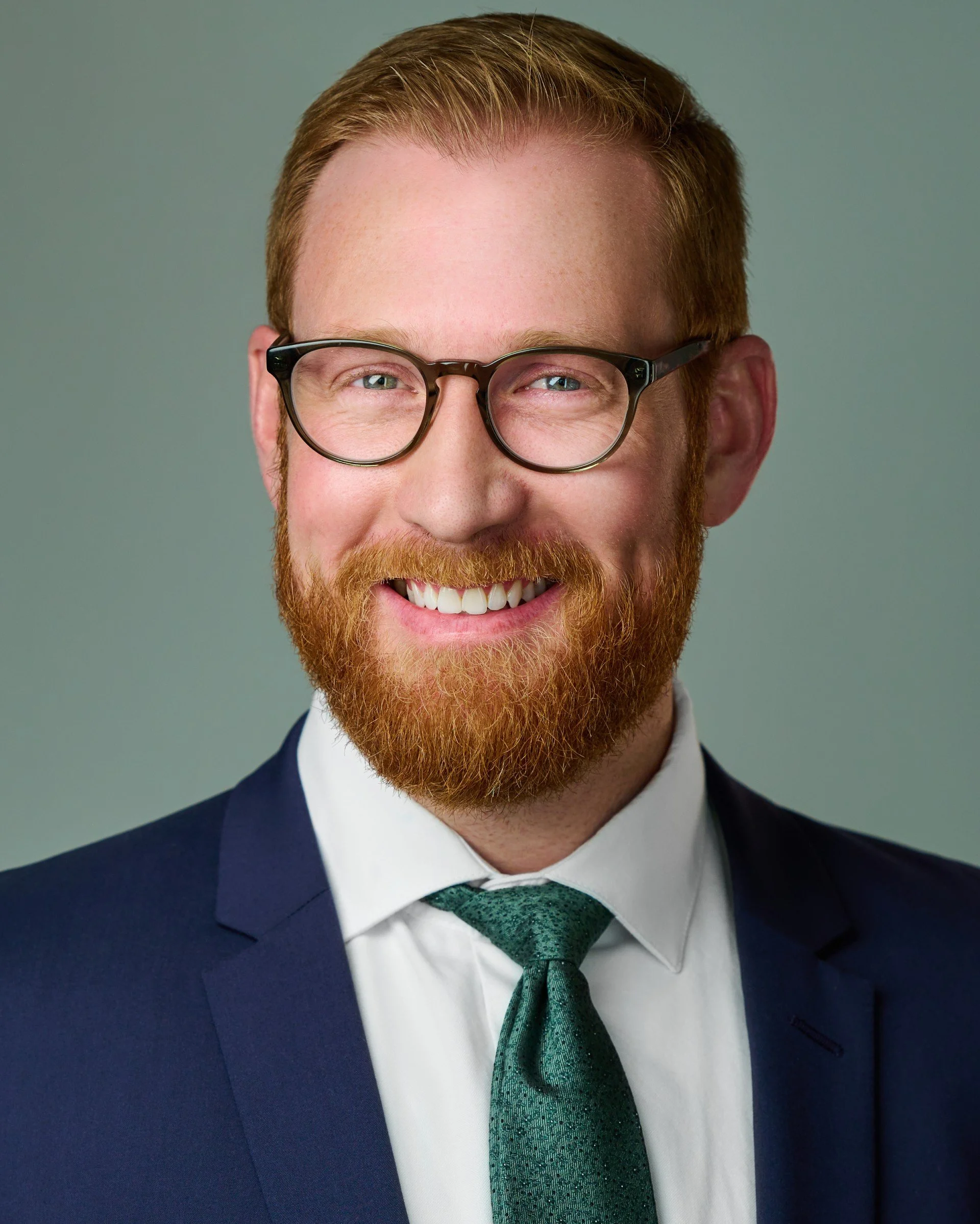 Close-up portrait of a man with red hair, a beard, glasses, wearing a suit, white shirt, and a green tie, smiling.