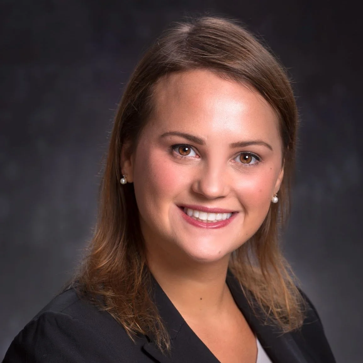 Professional headshot of a young woman with shoulder-length brown hair, wearing pearl earrings and a black blazer, smiling against a dark background.