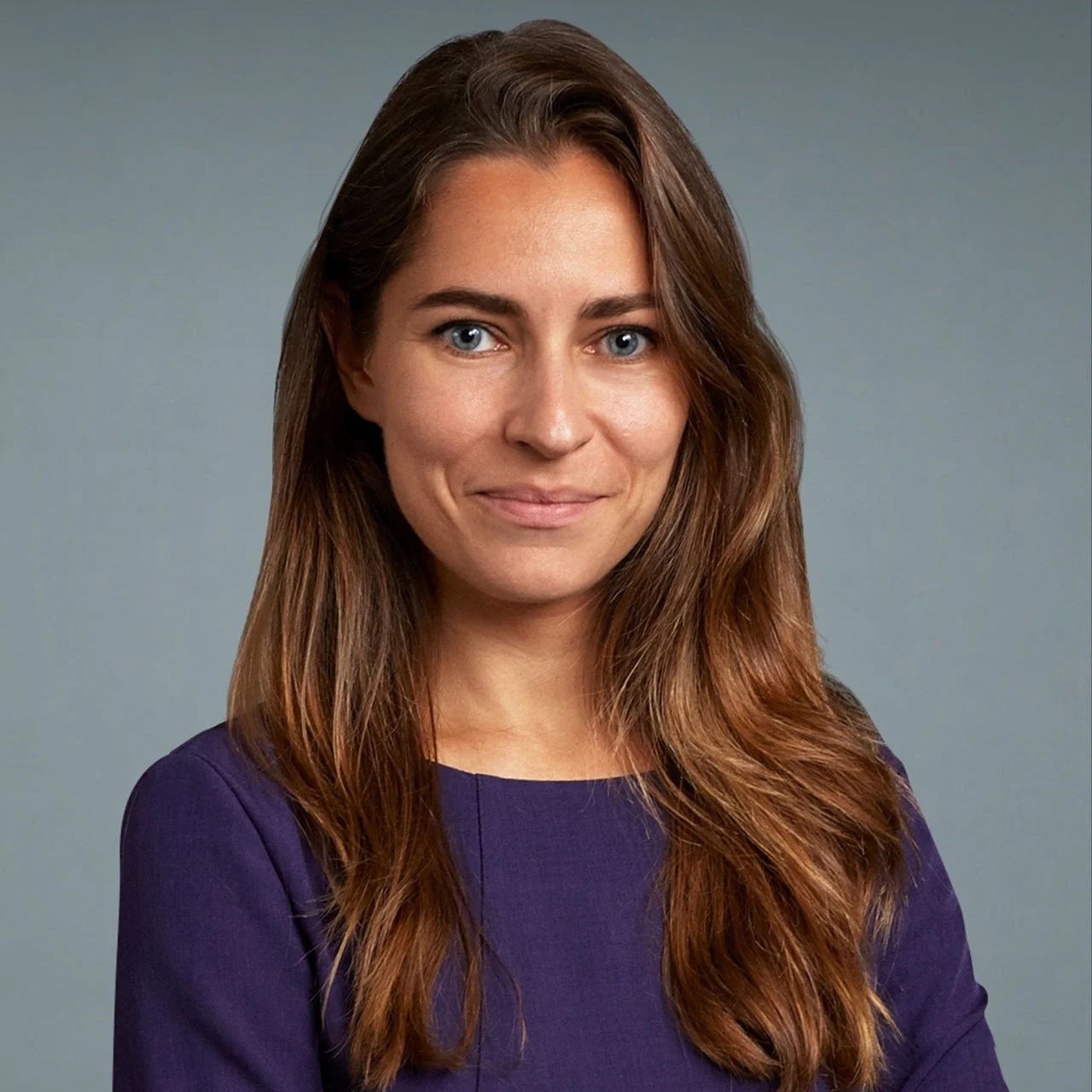 A woman with long, wavy brown hair and blue eyes wearing a purple top, smiling softly, against a gray background.