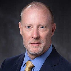 A middle-aged man with a bald head and gray beard, wearing a blue suit and a yellow tie, against a dark background.