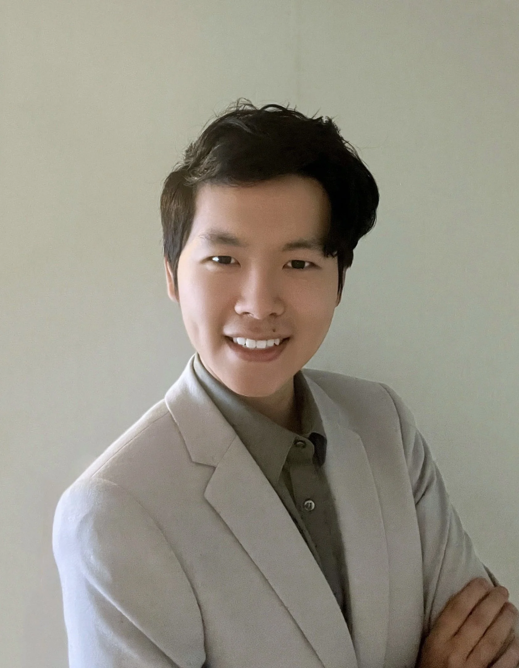 A young man with short dark hair, smiling and wearing a beige blazer over a dark shirt, standing against a plain light-colored background.