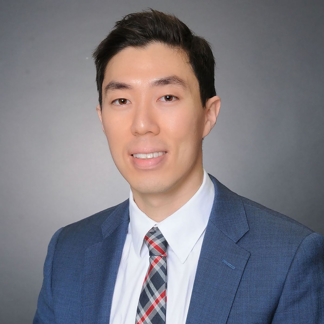 Professional portrait of a young man in a navy suit, white shirt, and checkered tie, smiling, against a plain gray background.