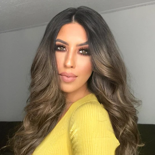 A woman with long, wavy brown hair wearing a yellow top, posing indoors.