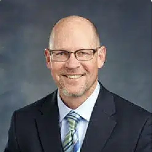 A professional headshot of a smiling man wearing a dark suit, light blue shirt, striped tie, and glasses with a dark background.