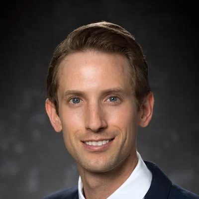 A professional headshot of a man with short brown hair, blue eyes, and a light complexion, smiling slightly, against a dark background.