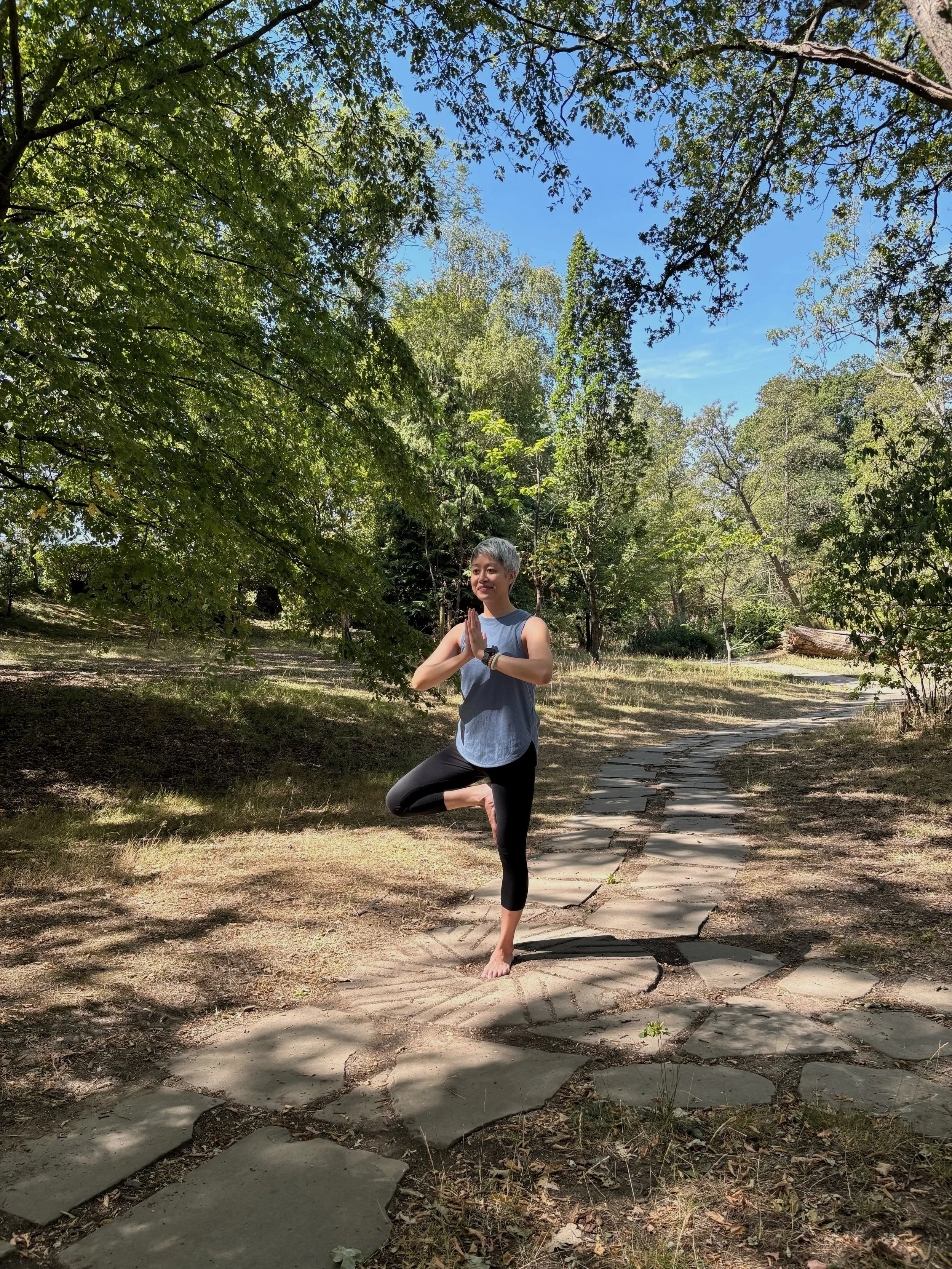 Ping standing in tree pose, with hands in prayer position, in a natural sunlit spot framed by trees .