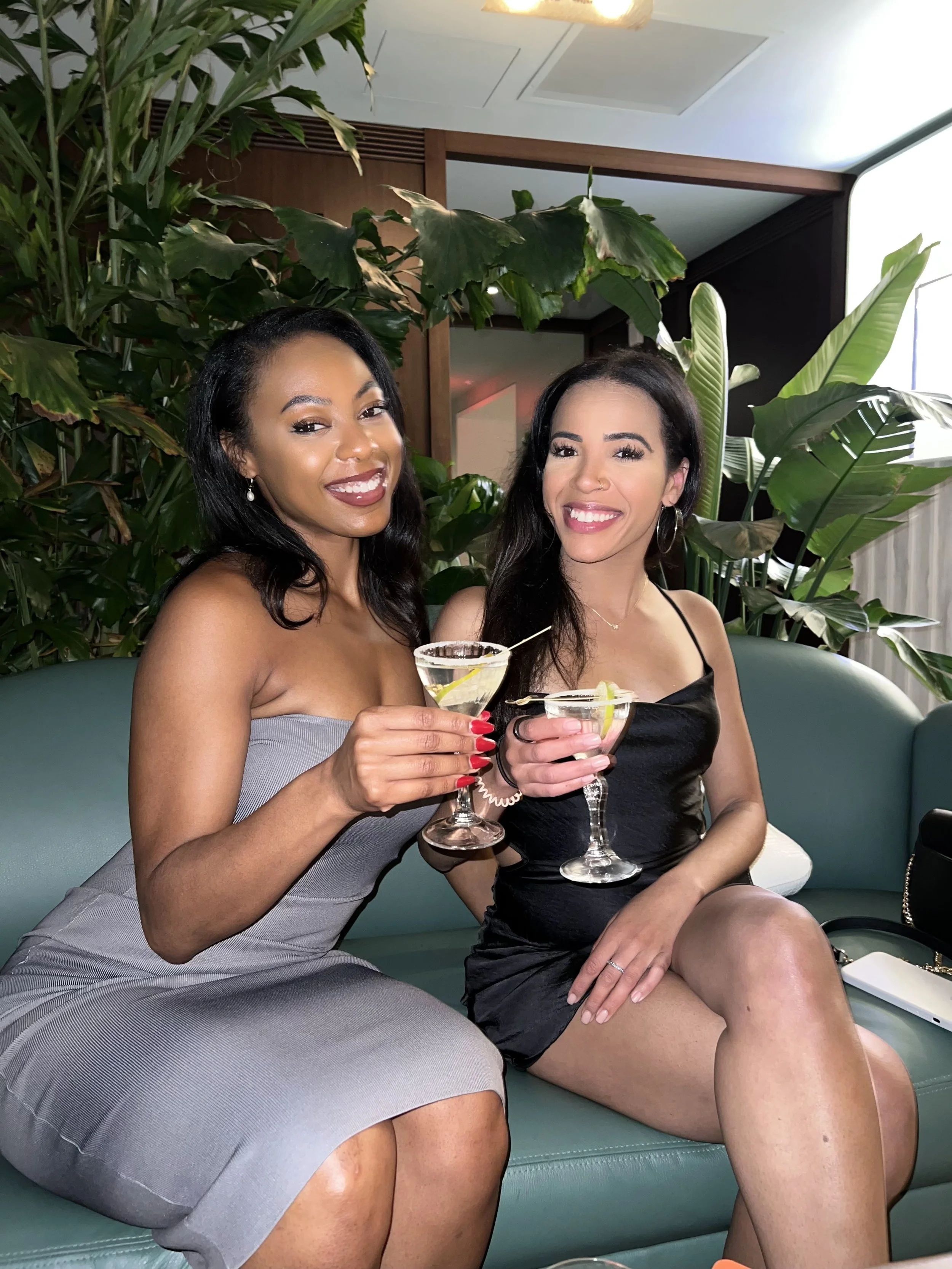 Two women sitting on a teal couch, smiling, holding cocktails garnished with lemon slices in a room with green plants and wood decor.